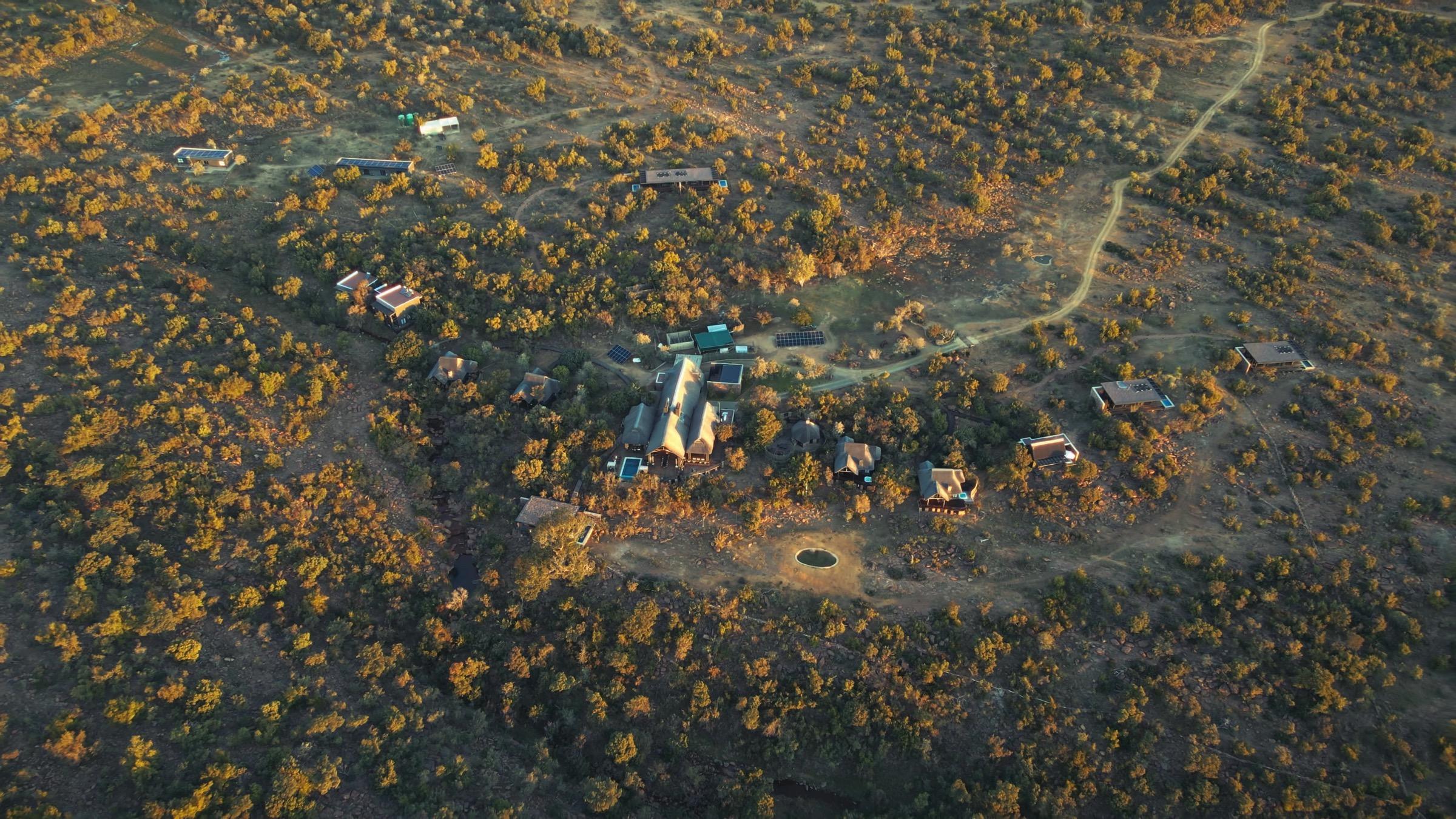 An aerial view of the whole lodge property at golden hour, several thatched buildings linked by paths and scattered across the bush with hills in the distance.
