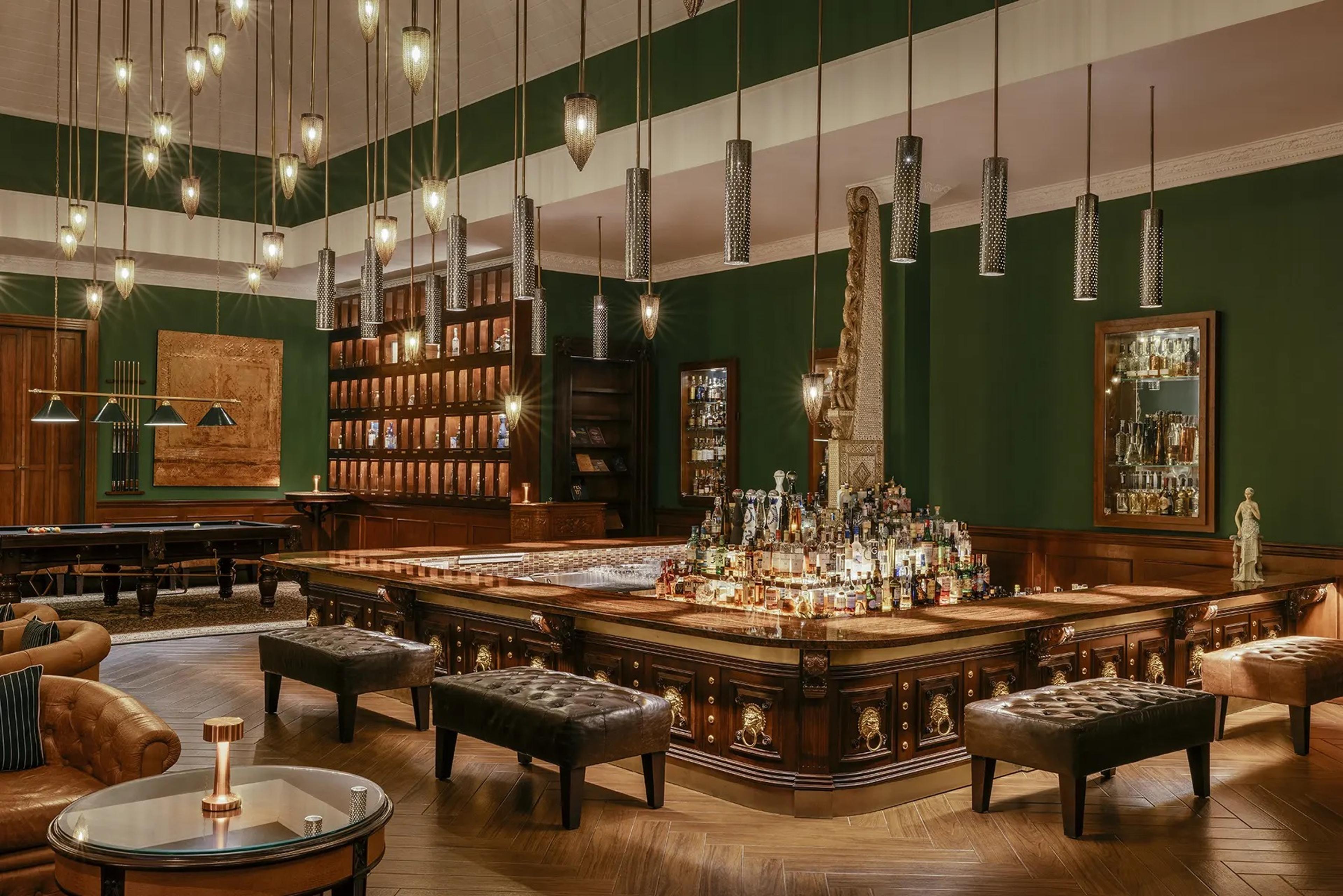 A dark green bar room with a billiard table, leather footstools around a central bar, and rows of hanging lights.