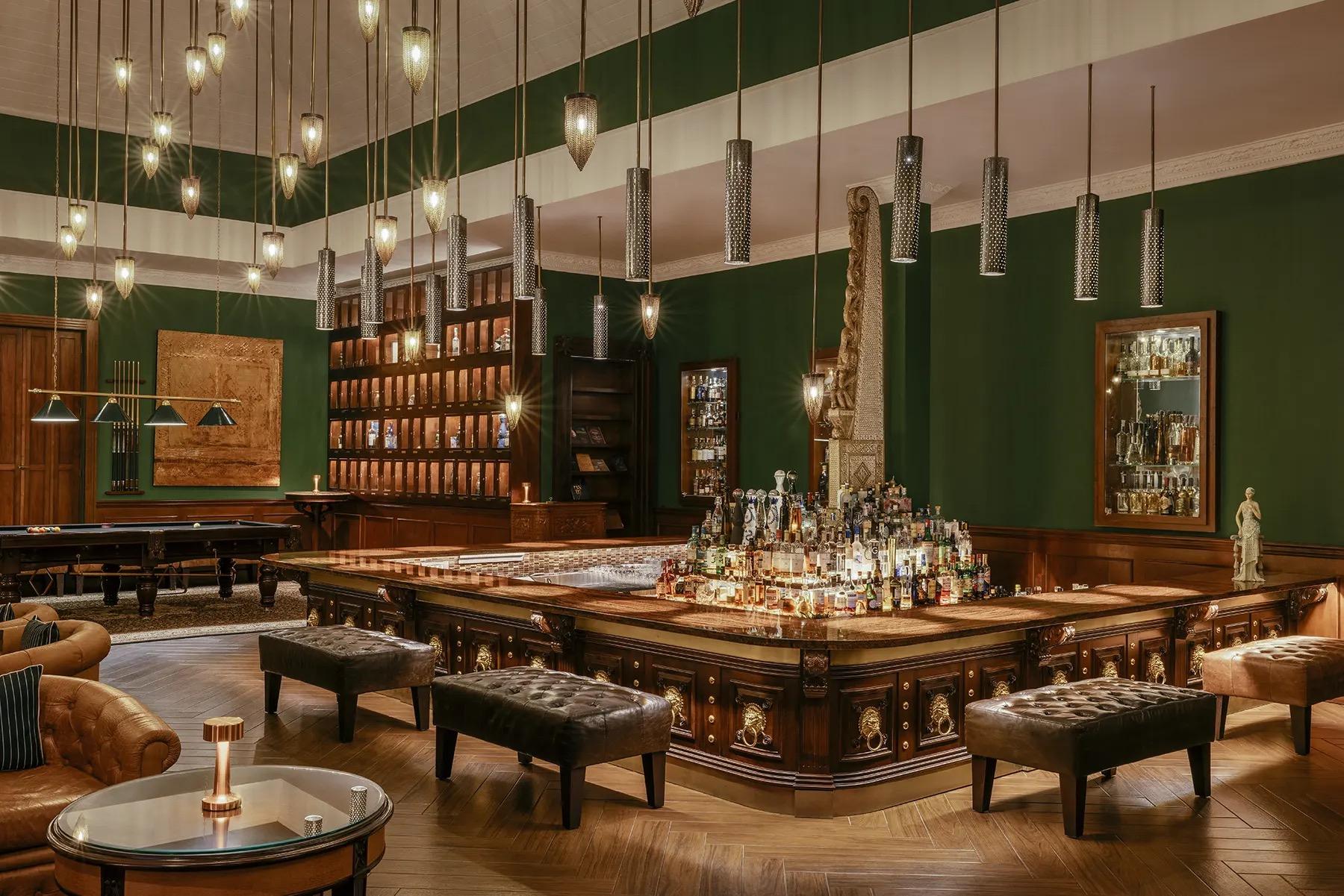 A dark green bar room with a billiard table, leather footstools around a central bar, and rows of hanging lights.