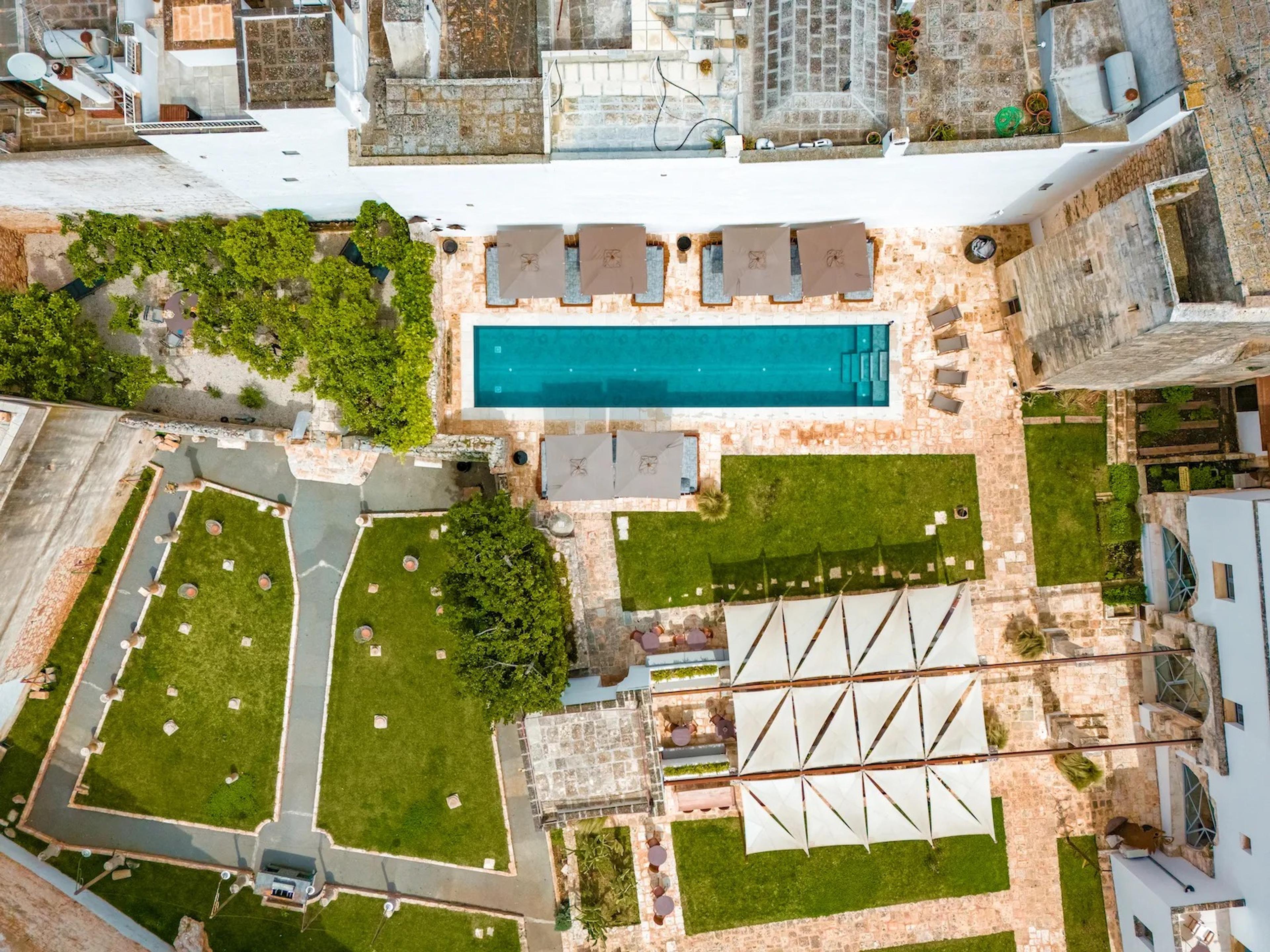 An aerial view of the hotel's courtyard, showing the pool, lawns, a sail-shaded dining area, and the surrounding stone buildings.