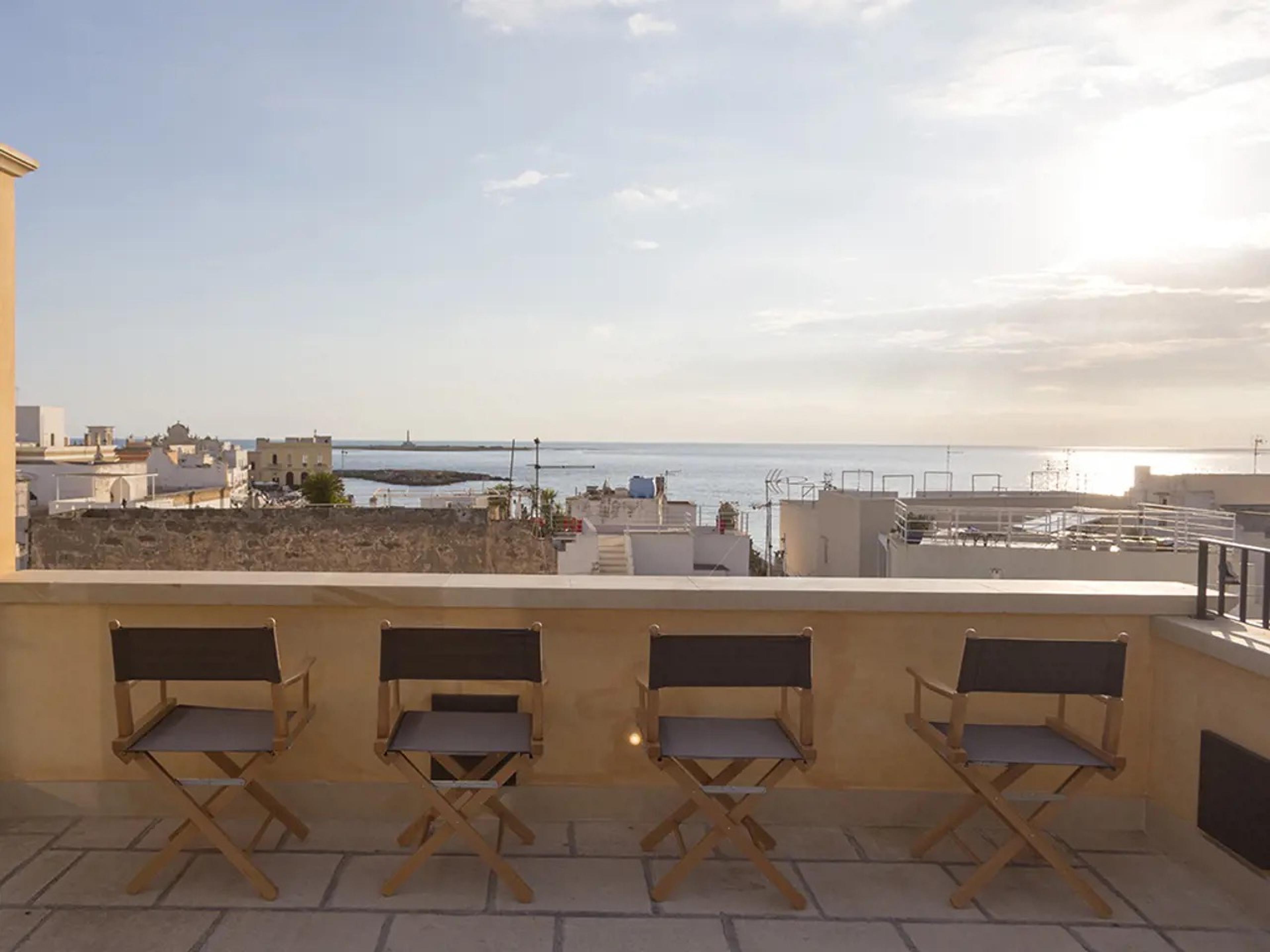 Four folding director's chairs lined along a rooftop parapet, facing an open sea view with the old town and harbour wall visible below.