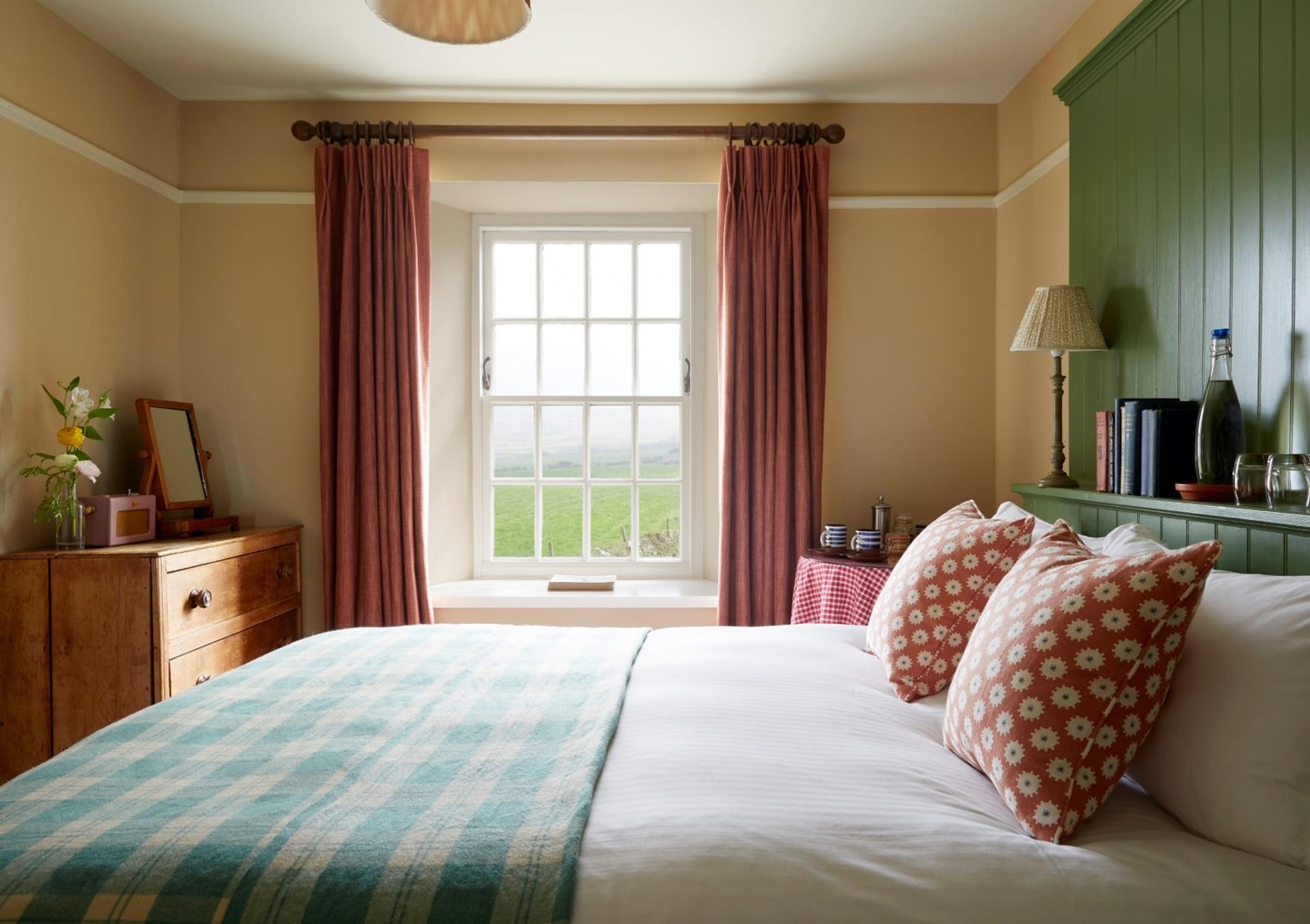 A bedroom with a window onto fields, a bed with a checked throw and patterned cushions, and a chest of drawers beside it.