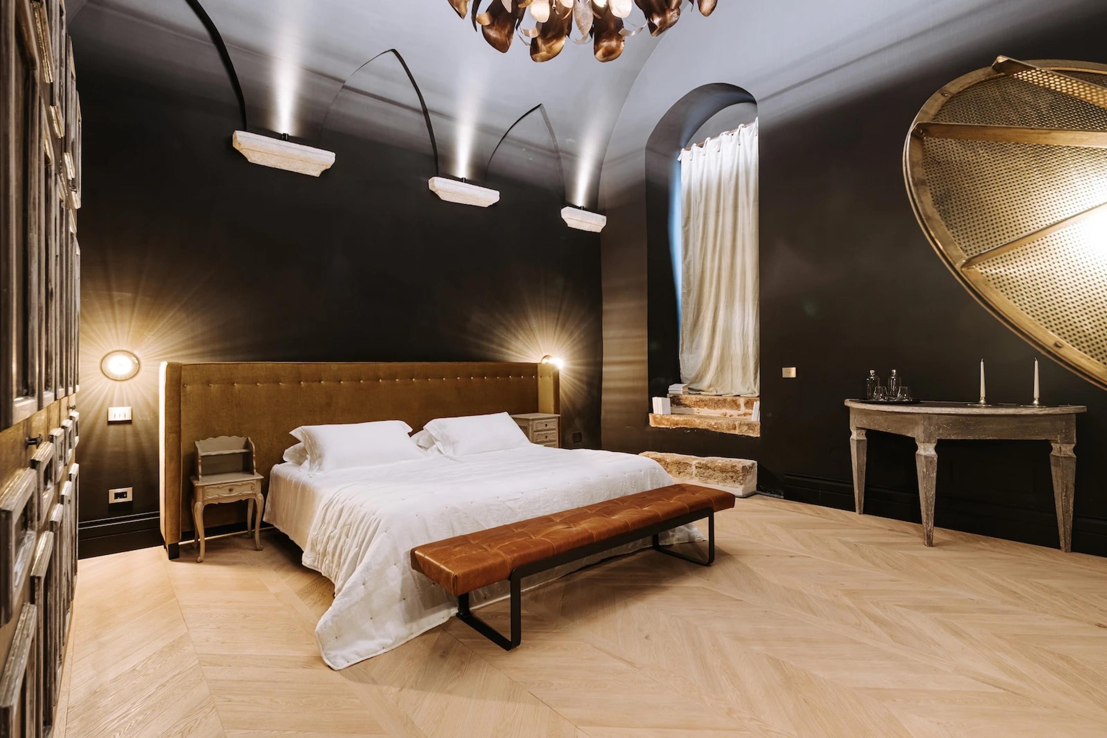 A dark-walled bedroom with a mustard velvet tufted headboard, a round gold mesh mirror, and a leather bench at the foot of the bed.