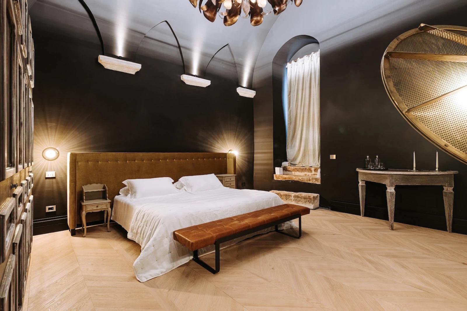 A dark-walled bedroom with a mustard velvet tufted headboard, a round gold mesh mirror, and a leather bench at the foot of the bed.