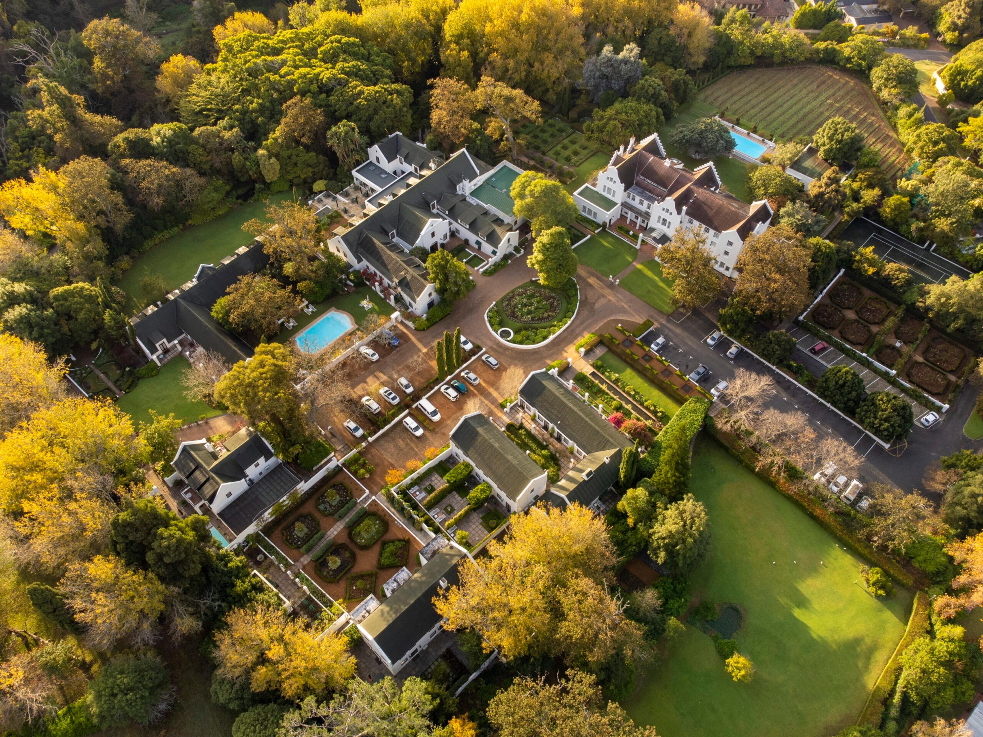 An aerial view of a hotel complex with pitched roofs, car parks, lawns, a tennis court and two swimming pools among tall trees.