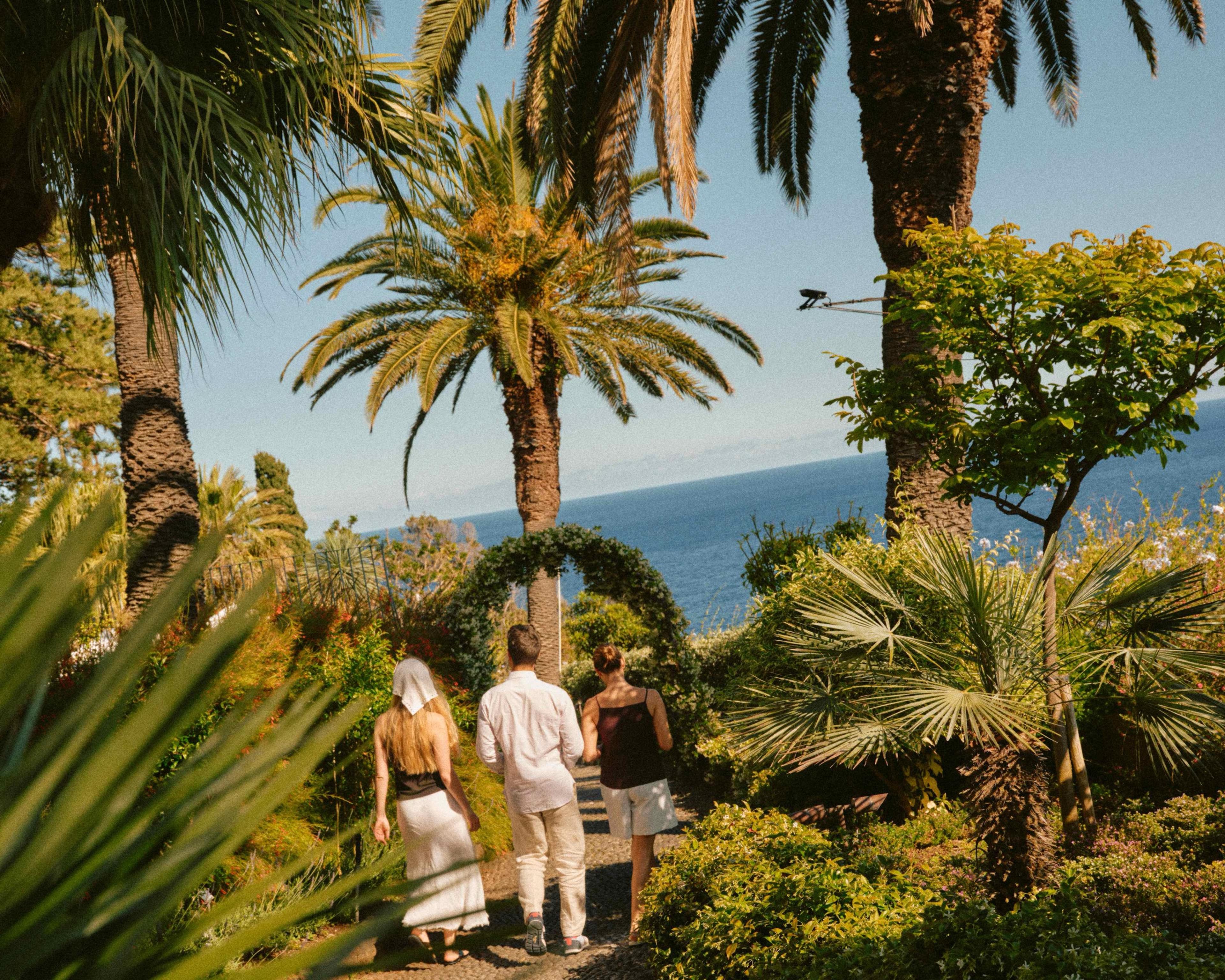 Three people walk away down a garden path lined with palm trees toward the sea, seen from behind.