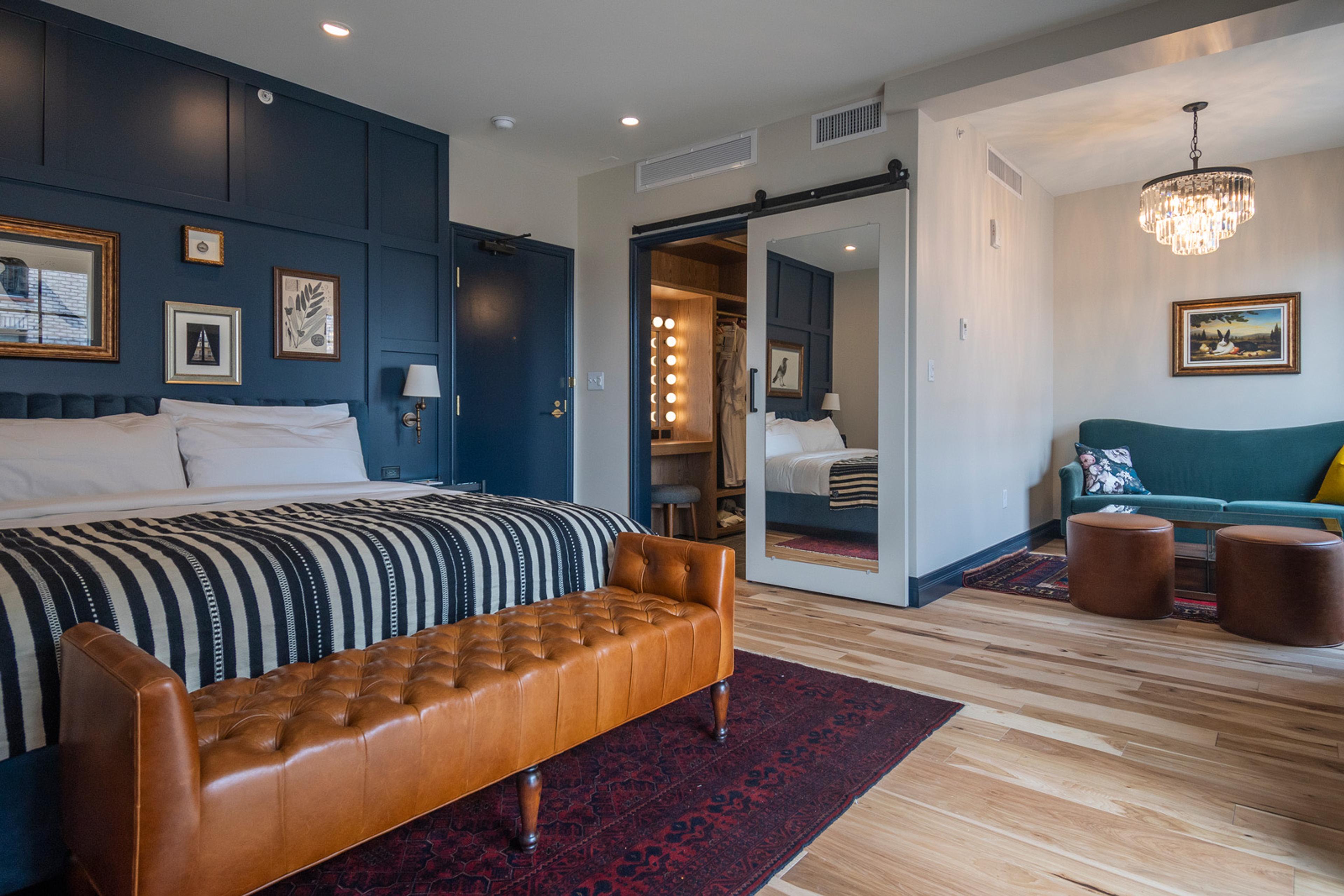 A room combining a bed with striped throw and leather bench, a wardrobe nook with a lit vanity mirror, and a teal sofa under a chandelier.