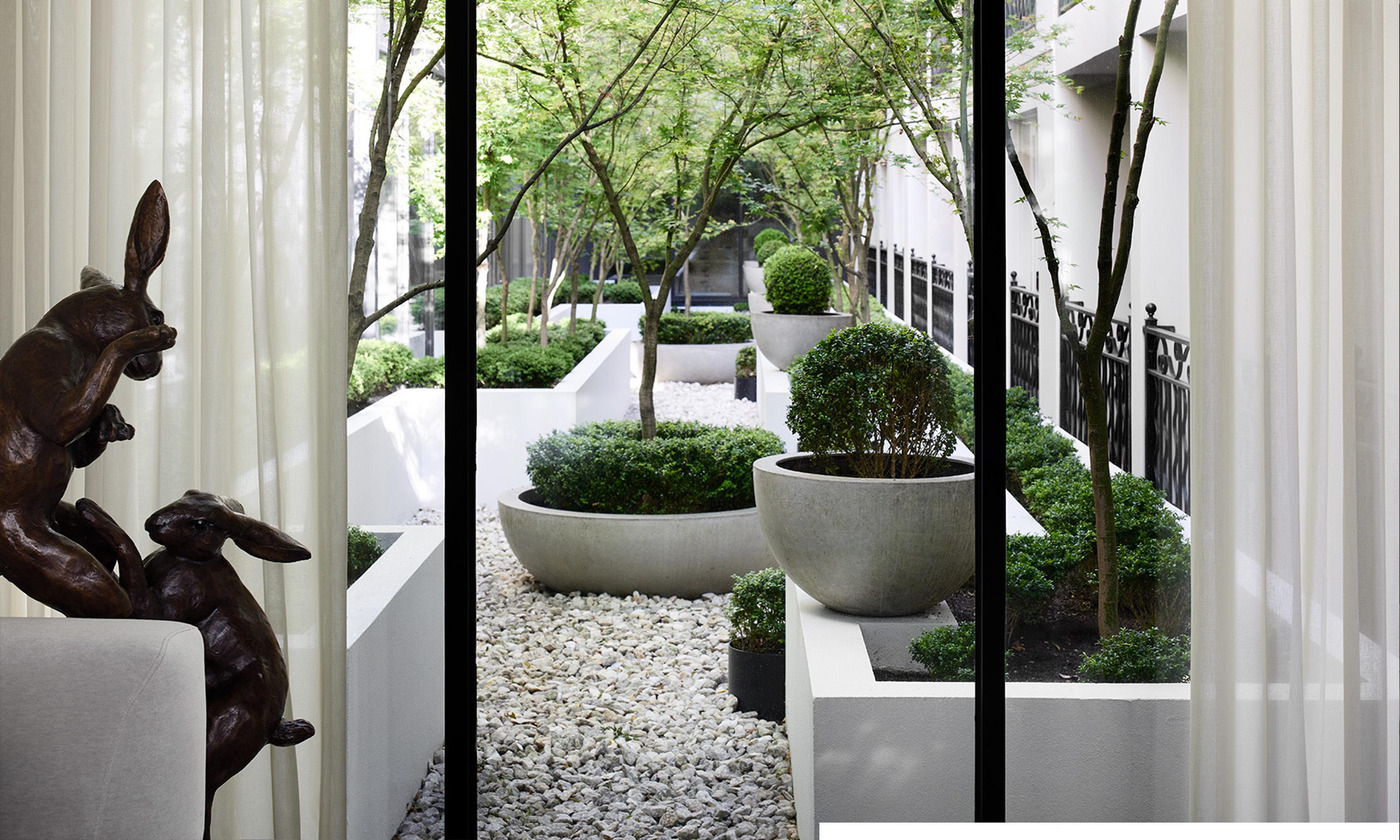 A paved courtyard seen through glass doors, with gravel, clipped shrubs in planters, trees overhead and a wrought iron fence.