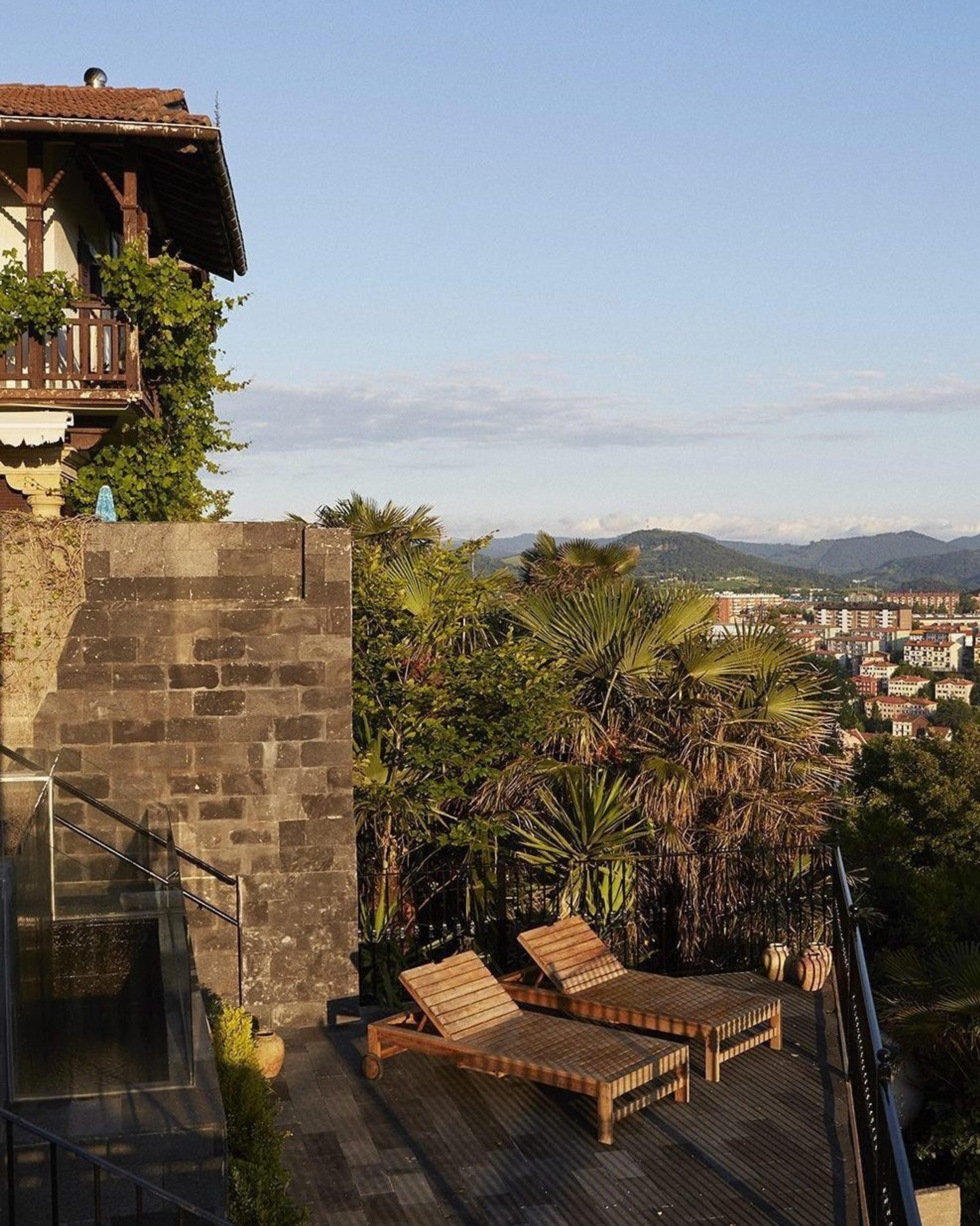 the corner of a stone and white-rendered building with a tiled roof and balcony, palm trees and two loungers on the terrace below, the town spread out beyond