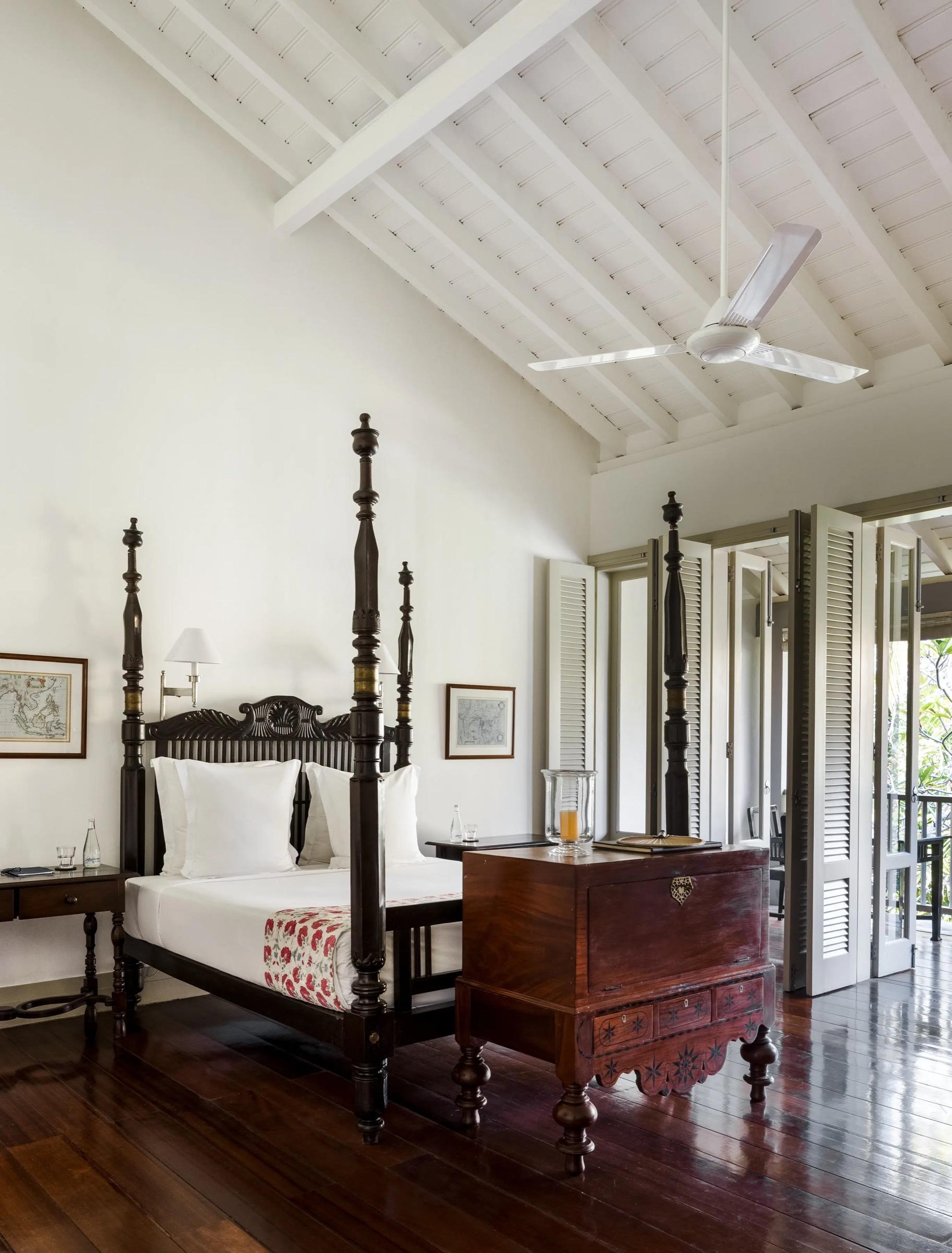 A high-ceilinged bedroom with a dark four poster bed, a wooden trunk at its foot, and louvred doors open to a balcony.