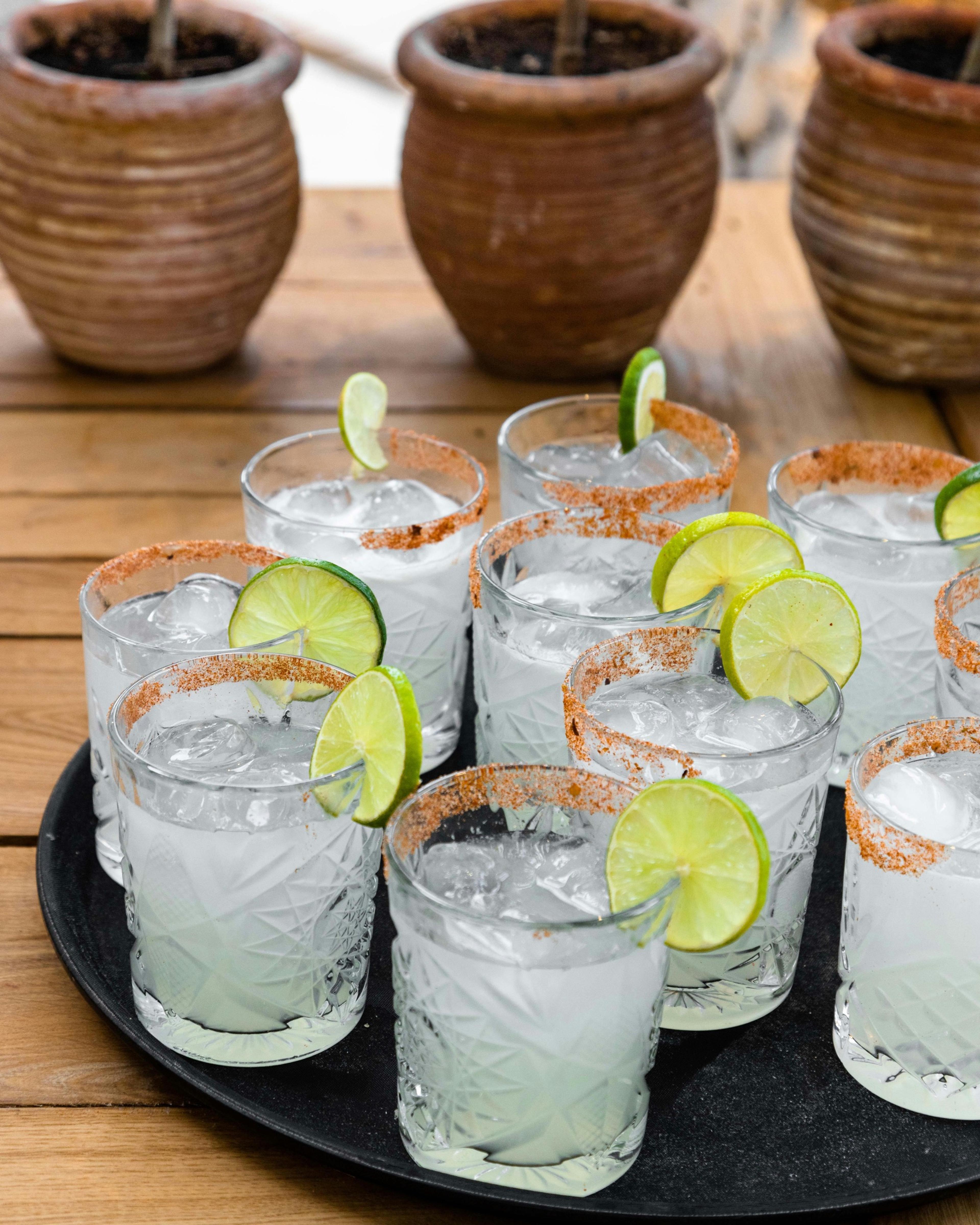 Ten iced cocktails with chilli salt rims and lime wheels are arranged on a black tray on a wooden table.
