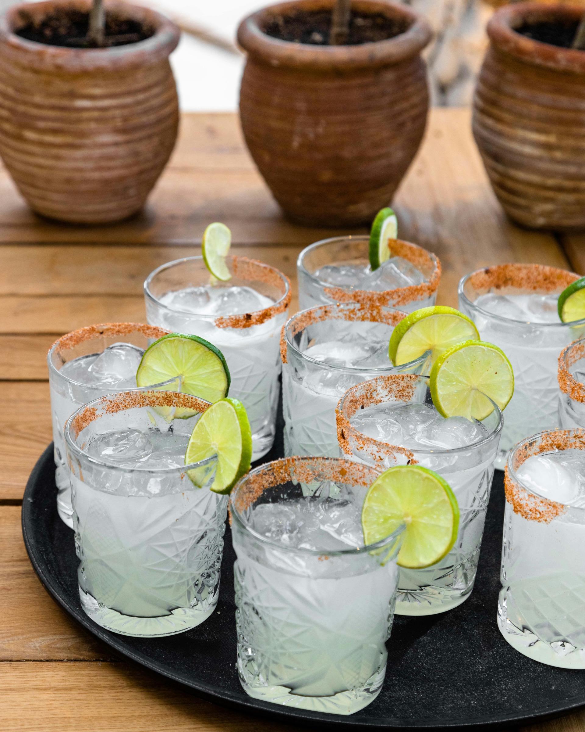Ten iced cocktails with chilli salt rims and lime wheels are arranged on a black tray on a wooden table.