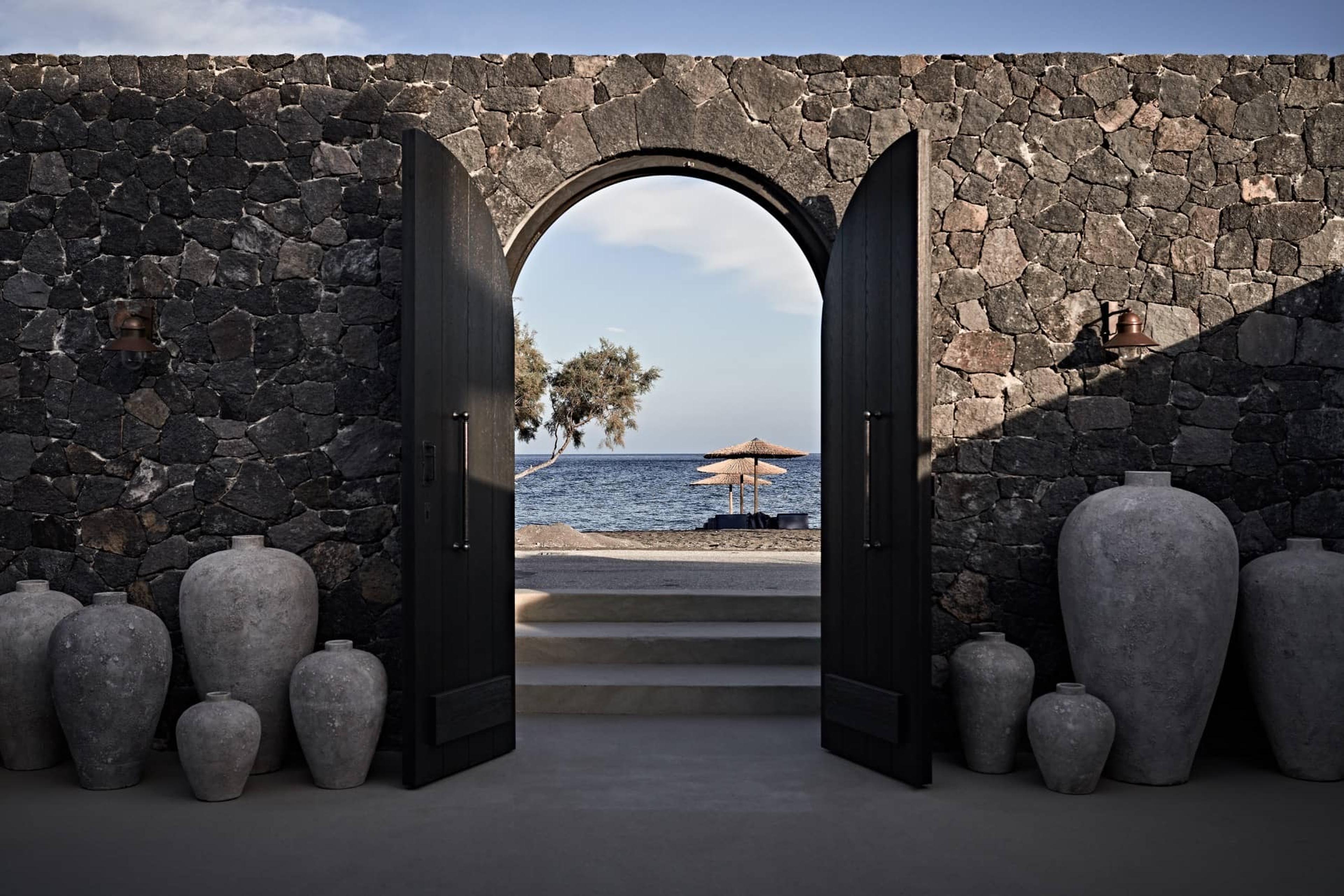 A stone archway in a thick wall, flanked by large ceramic pots, open to a view of a tree, a beach umbrella and the sea.