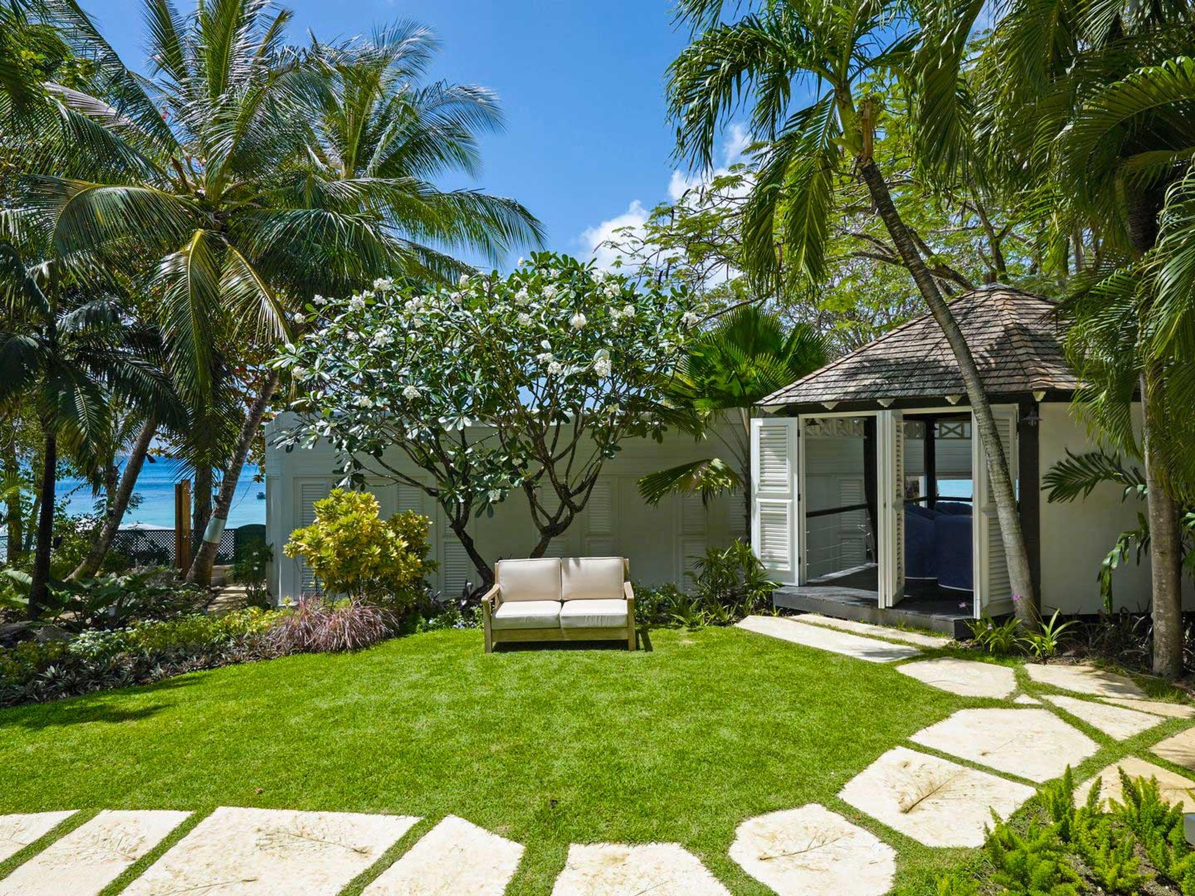 A tropical garden with a flowering frangipani tree, a lawn, an outdoor sofa and a white shuttered garden building among palm trees, the sea just visible beyond