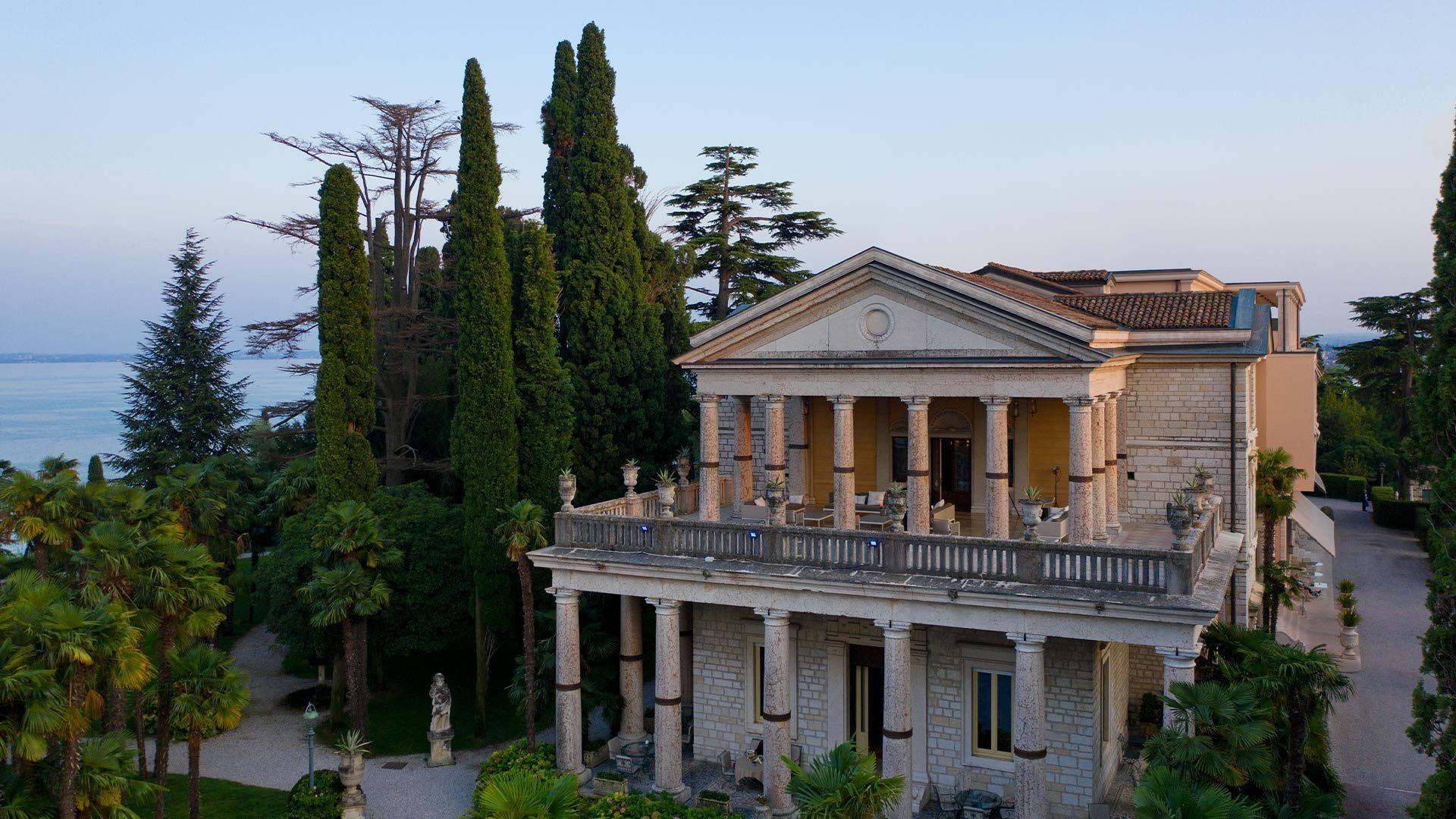 The neoclassical facade of a villa with columned balconies, seen through palm trees and cypresses.