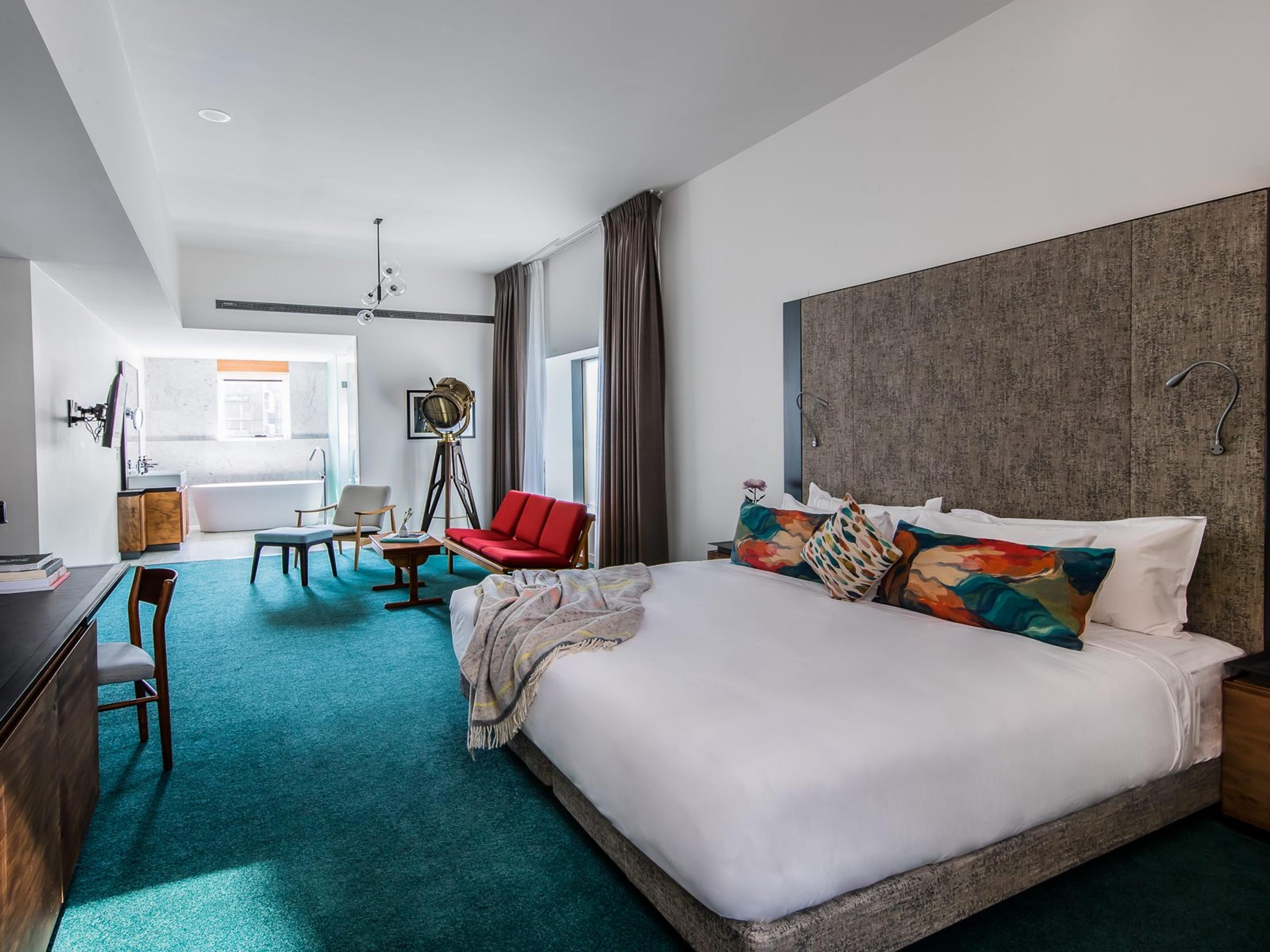 A loft suite with teal carpet, a bed with colourful cushions, a brass spotlight lamp, a red bench, and a freestanding bathtub in an open bathroom beyond.