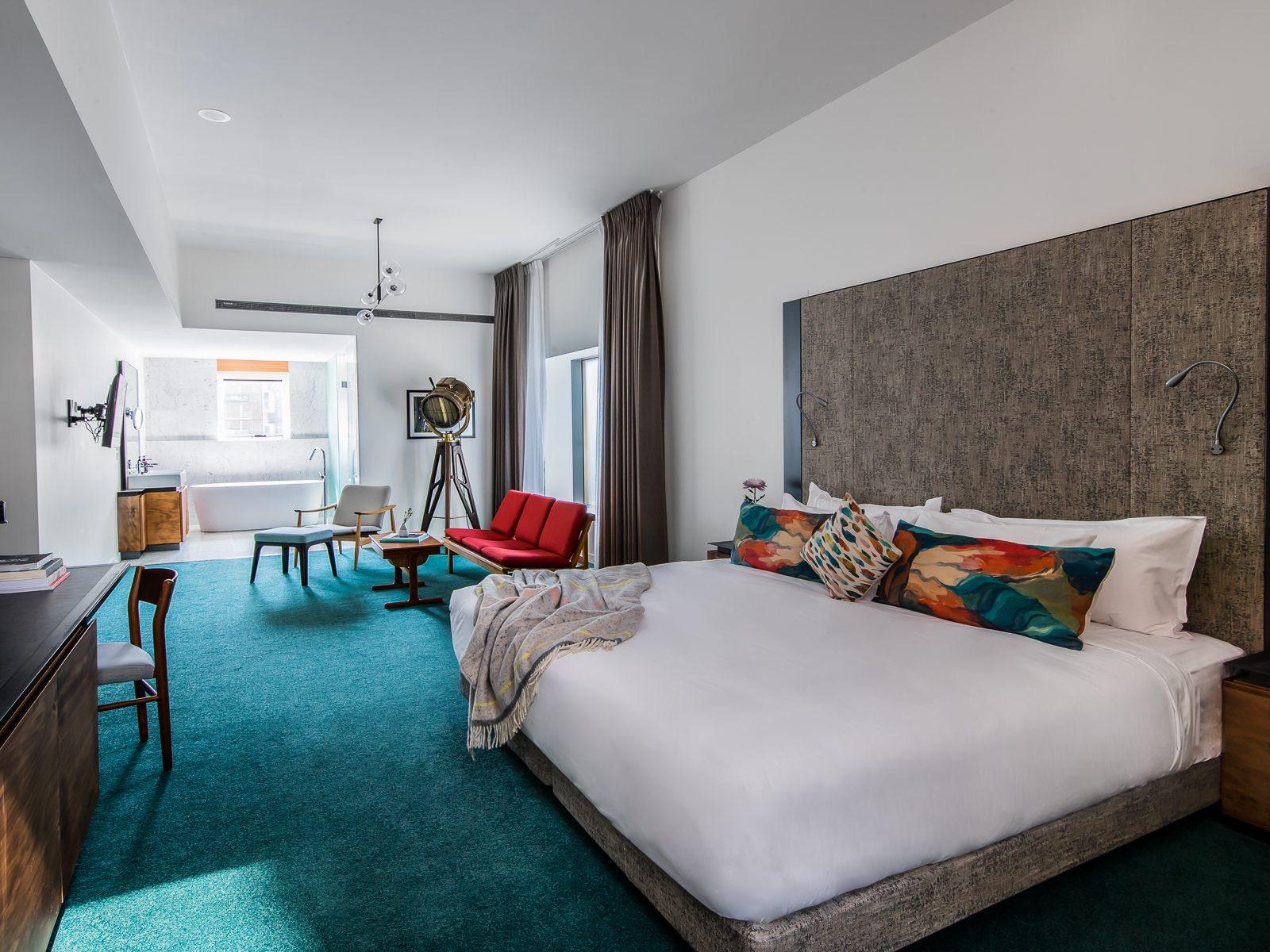 A loft suite with teal carpet, a bed with colourful cushions, a brass spotlight lamp, a red bench, and a freestanding bathtub in an open bathroom beyond.