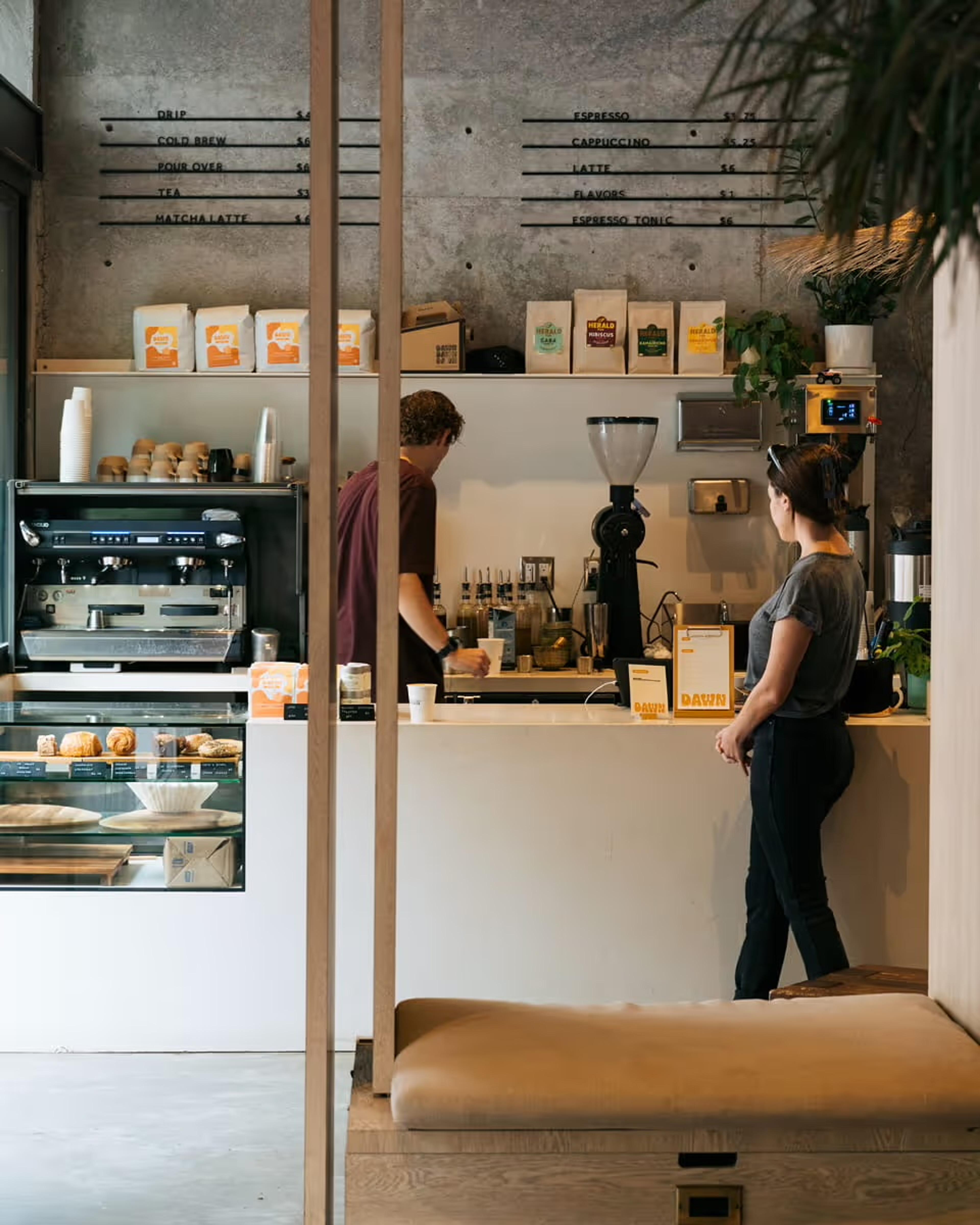 Inside the Dawn cafe, a barista works at the espresso machine while a customer waits at the counter, both seen from the side with their faces turned away.