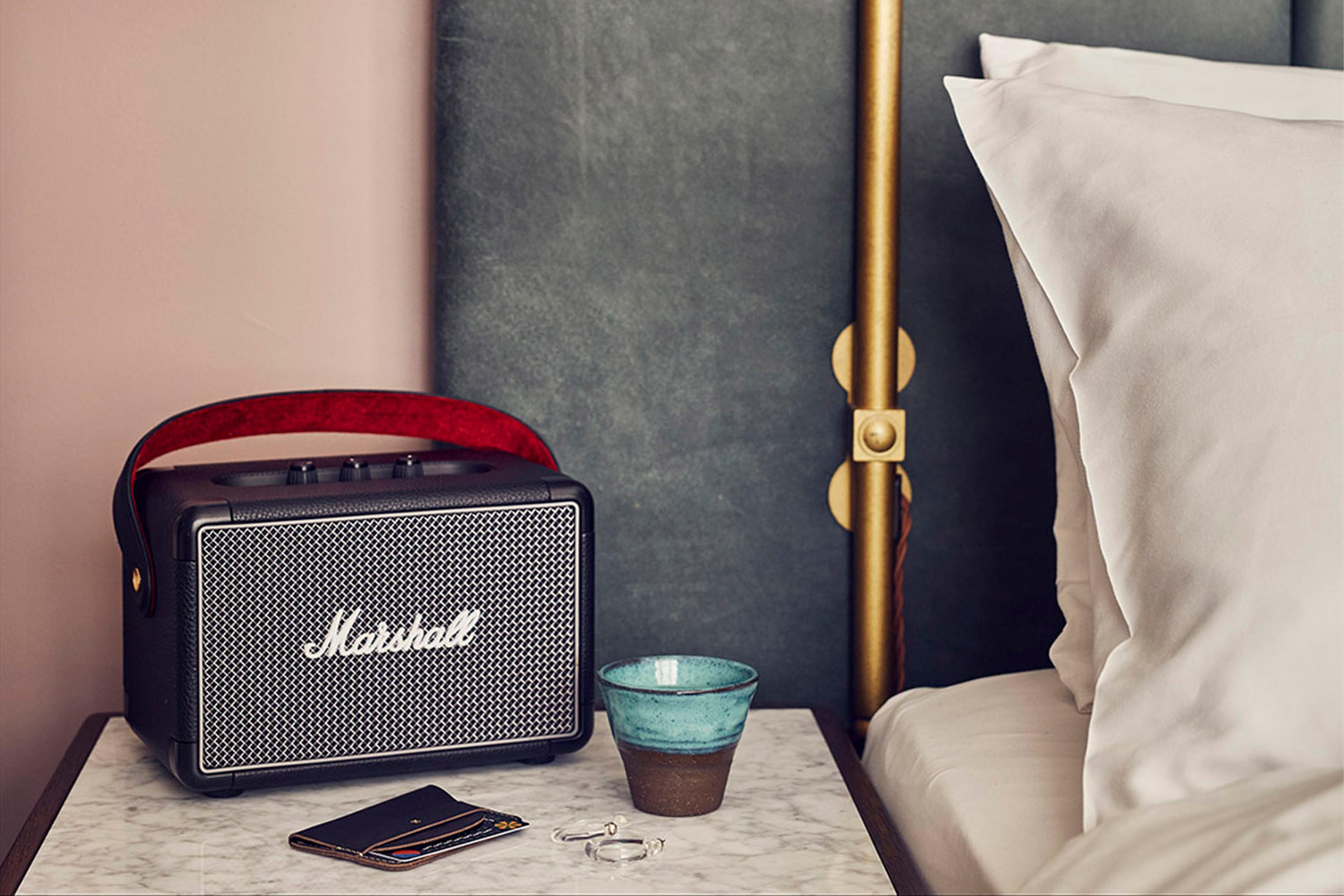 A black Marshall speaker, a teal ceramic cup, and a wallet on a marble nightstand beside a bed with white pillows and a brass lamp pole.