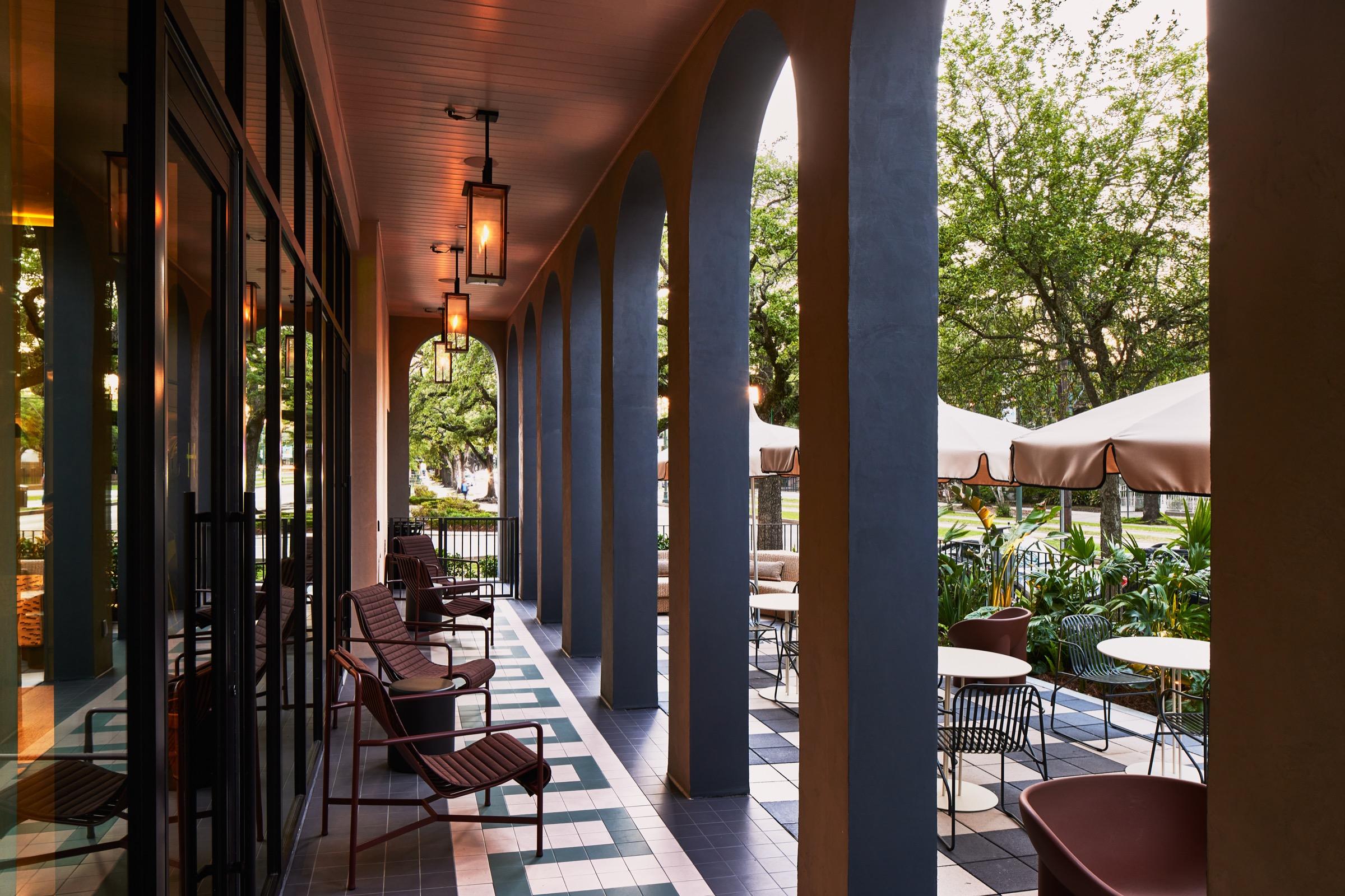 A colonnade of arched columns along a tiled terrace, with metal chairs and round tables under cream parasols and trees beyond.