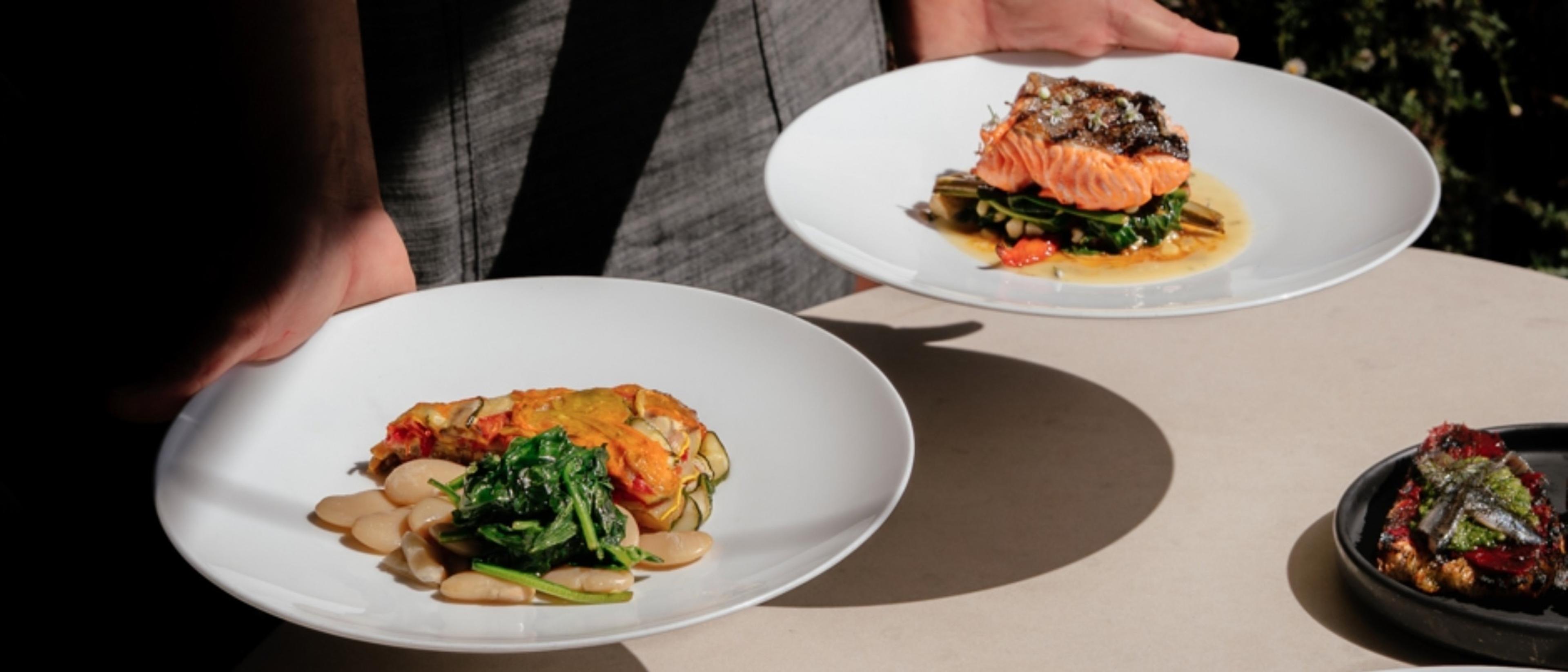 A person's hands holding two white plates of plated food, one with salmon and greens and one with a vegetable tart, on an outdoor table in sunlight.