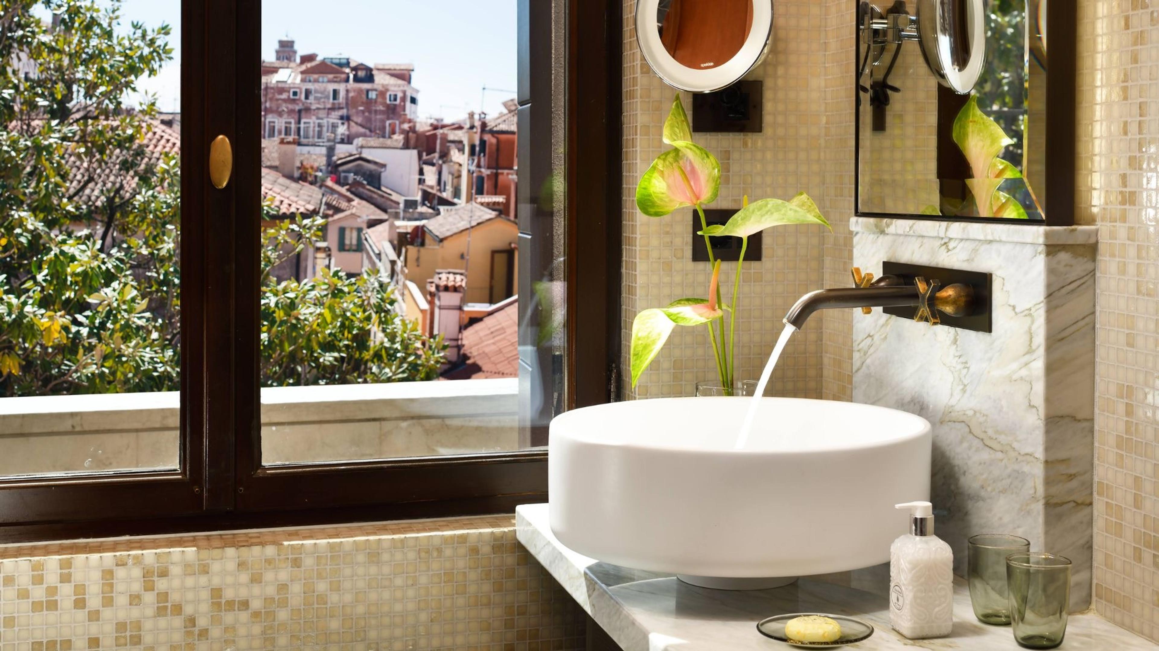 A bathroom sink with water running from the tap, a potted plant beside it and an open window looking over the tiled rooftops of Venice.