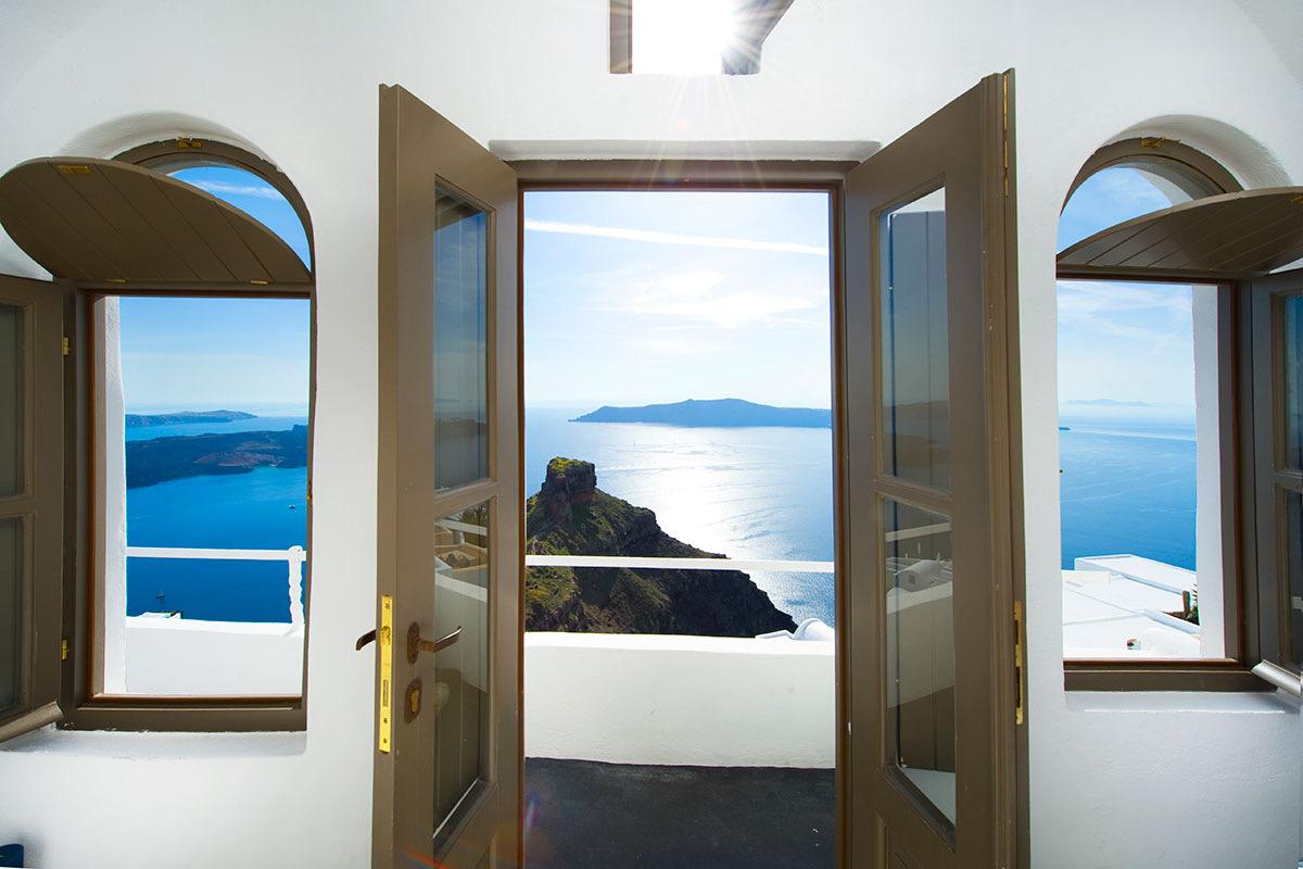 Through open double doors and a pair of arched windows, the view across the caldera sea to a rocky islet and a distant island.