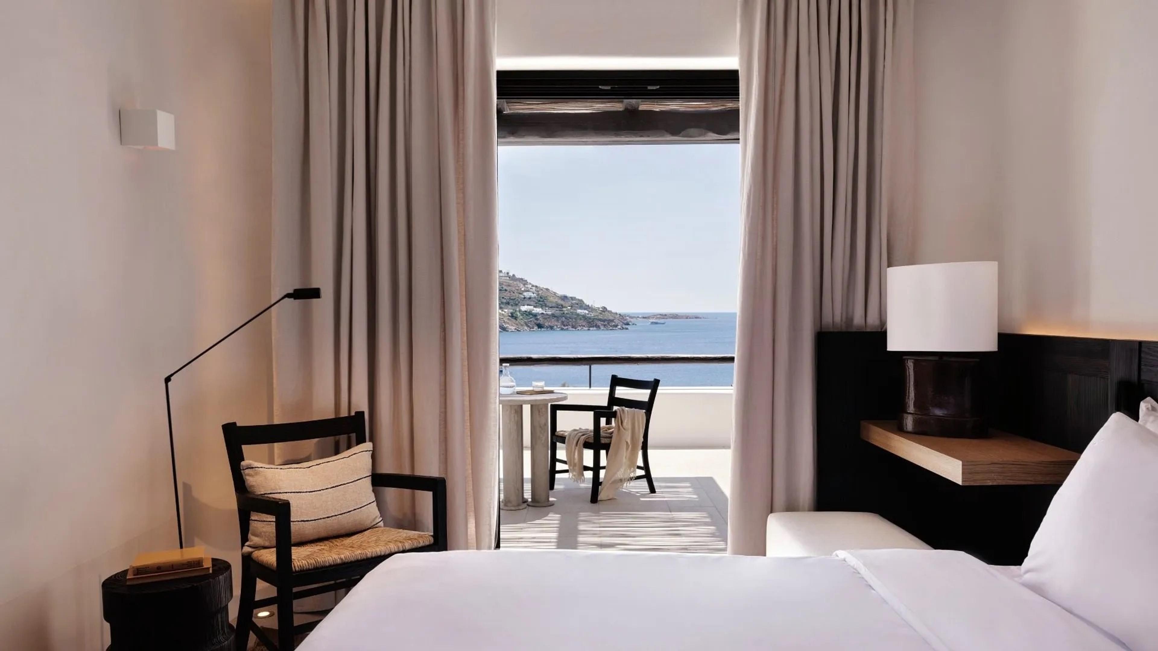 A bedroom with a white bed in the foreground, its curtains drawn back to a terrace with a table, two chairs and the sea beyond.