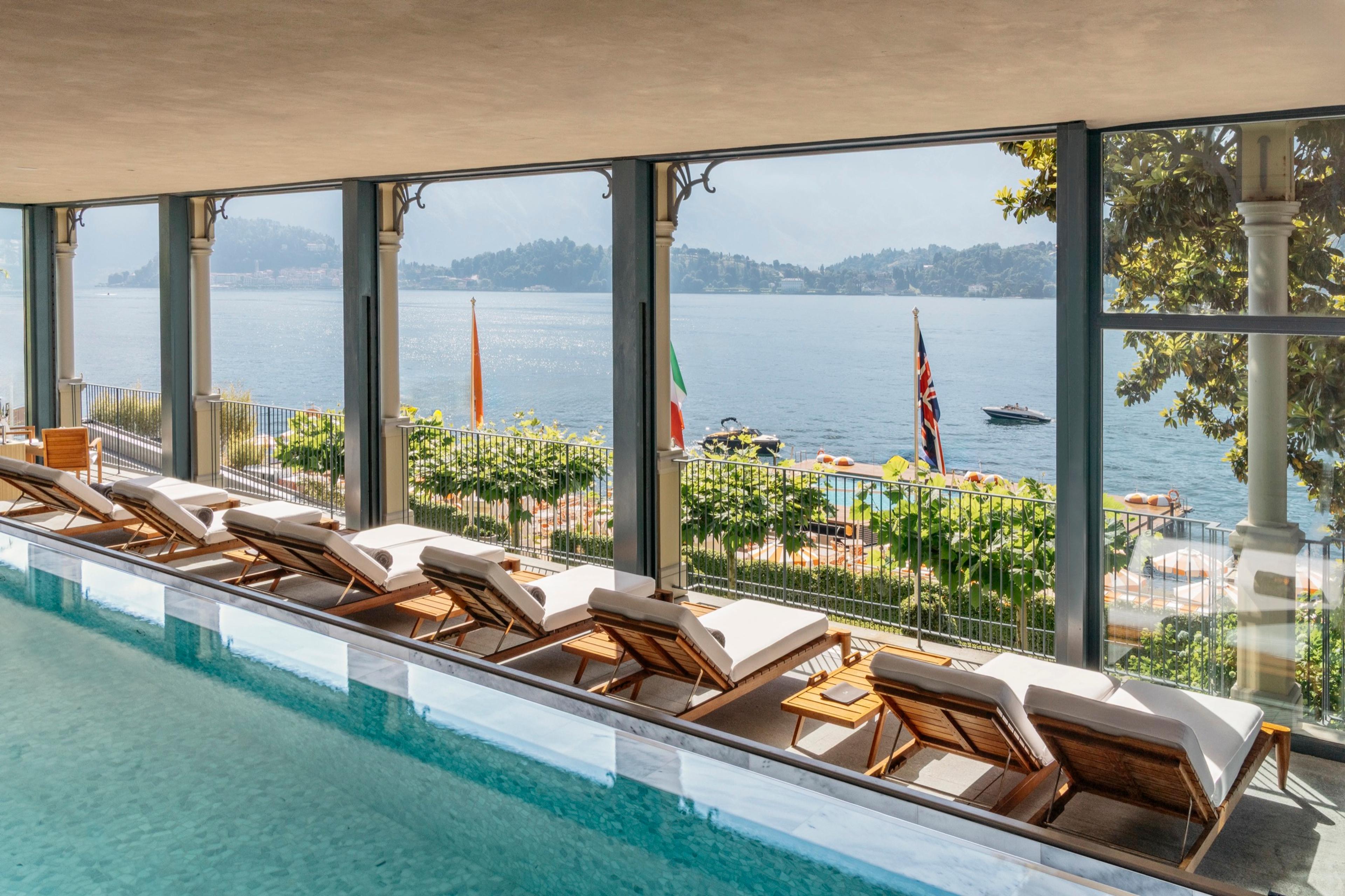 Wooden sun loungers line an indoor infinity pool, with glass doors open to a lake view, flags and moored boats.