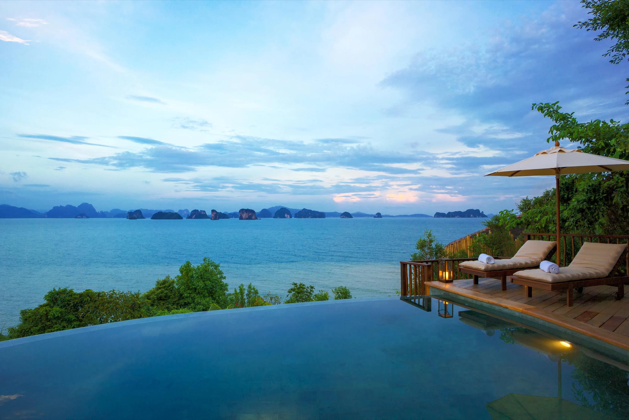 An infinity pool beside two cushioned loungers under a shaded umbrella, overlooking a bay scattered with islands at dusk.