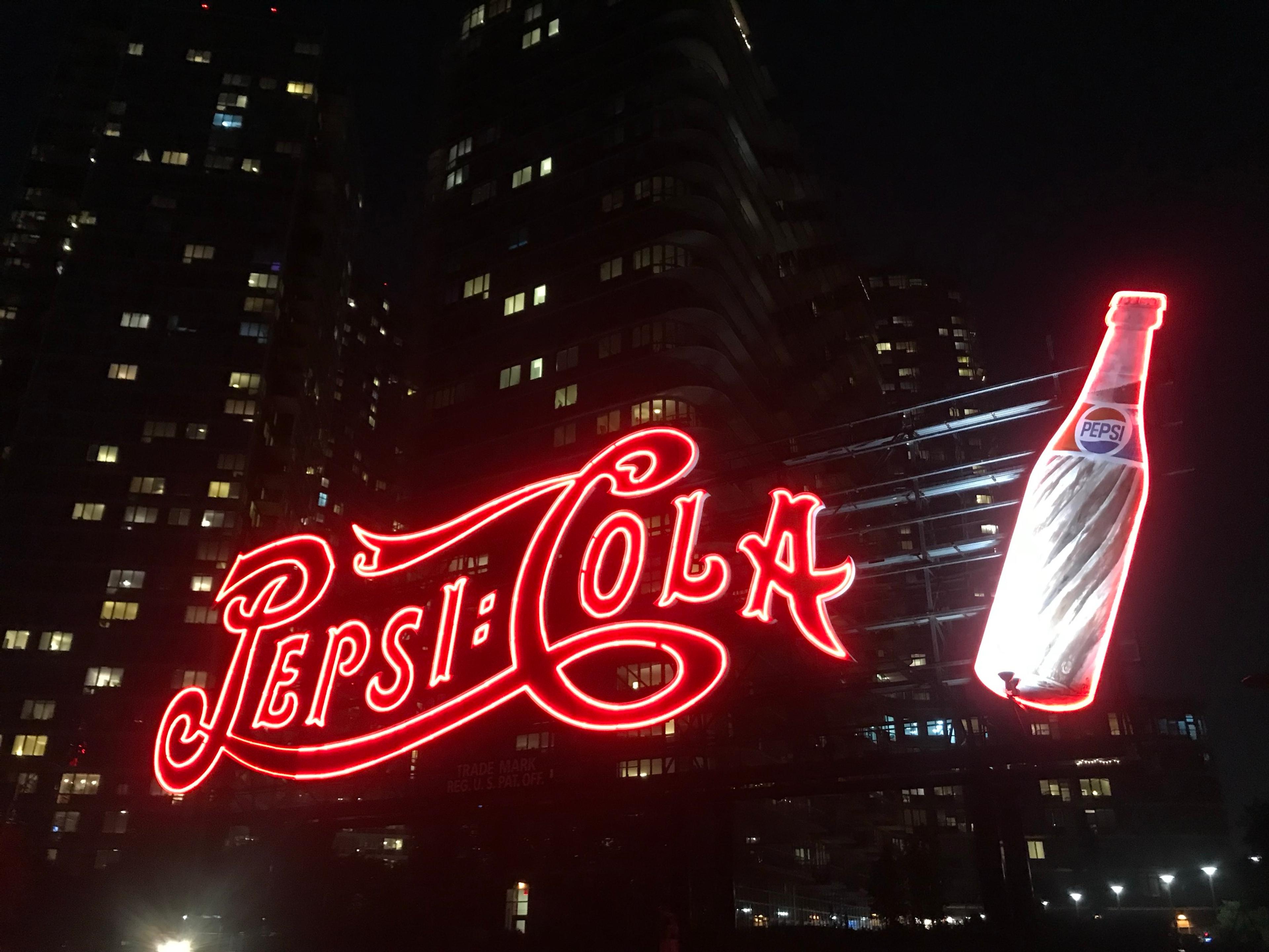 The red neon Pepsi-Cola sign at night on the Long Island City waterfront, apartment towers lit up behind it