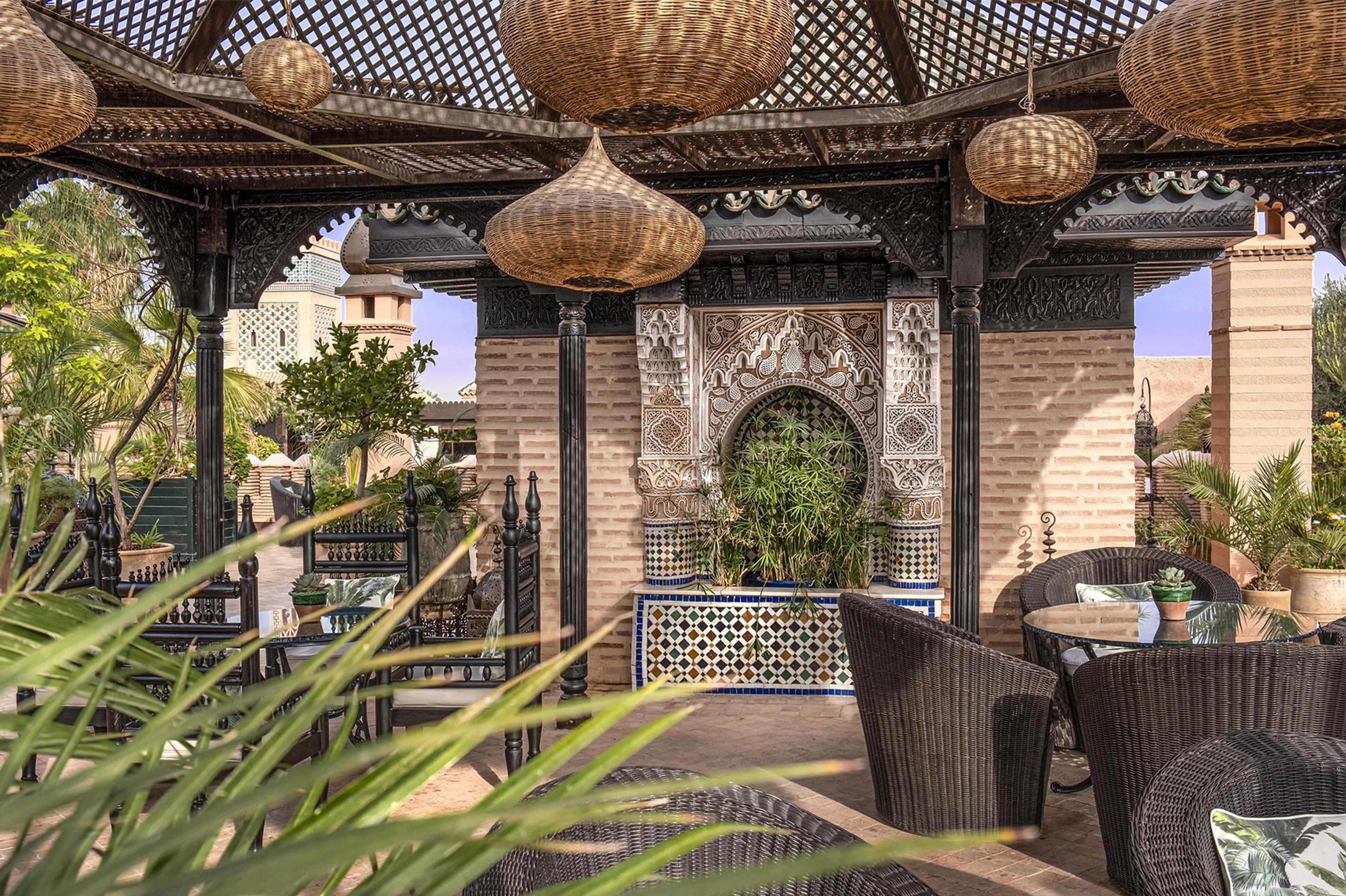 A covered garden pavilion with woven lantern shades hanging from a lattice roof, wicker chairs, and a tiled archway framing a planted niche.