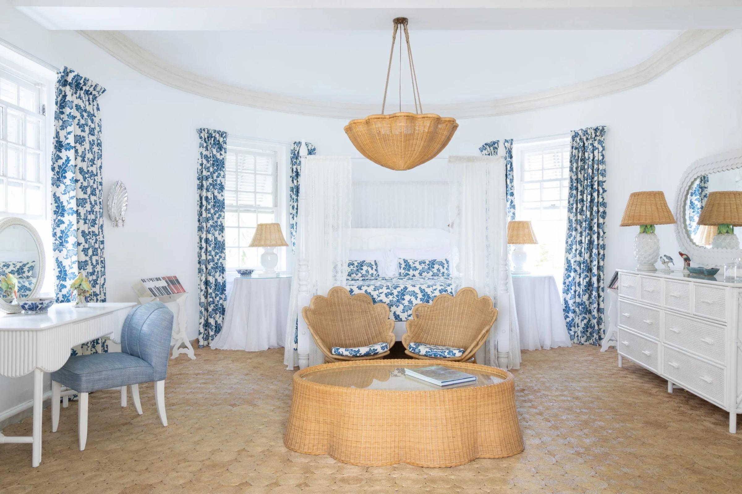 a bedroom with a white four-poster canopy bed dressed in blue floral linen, wicker armchairs and a round wicker coffee table