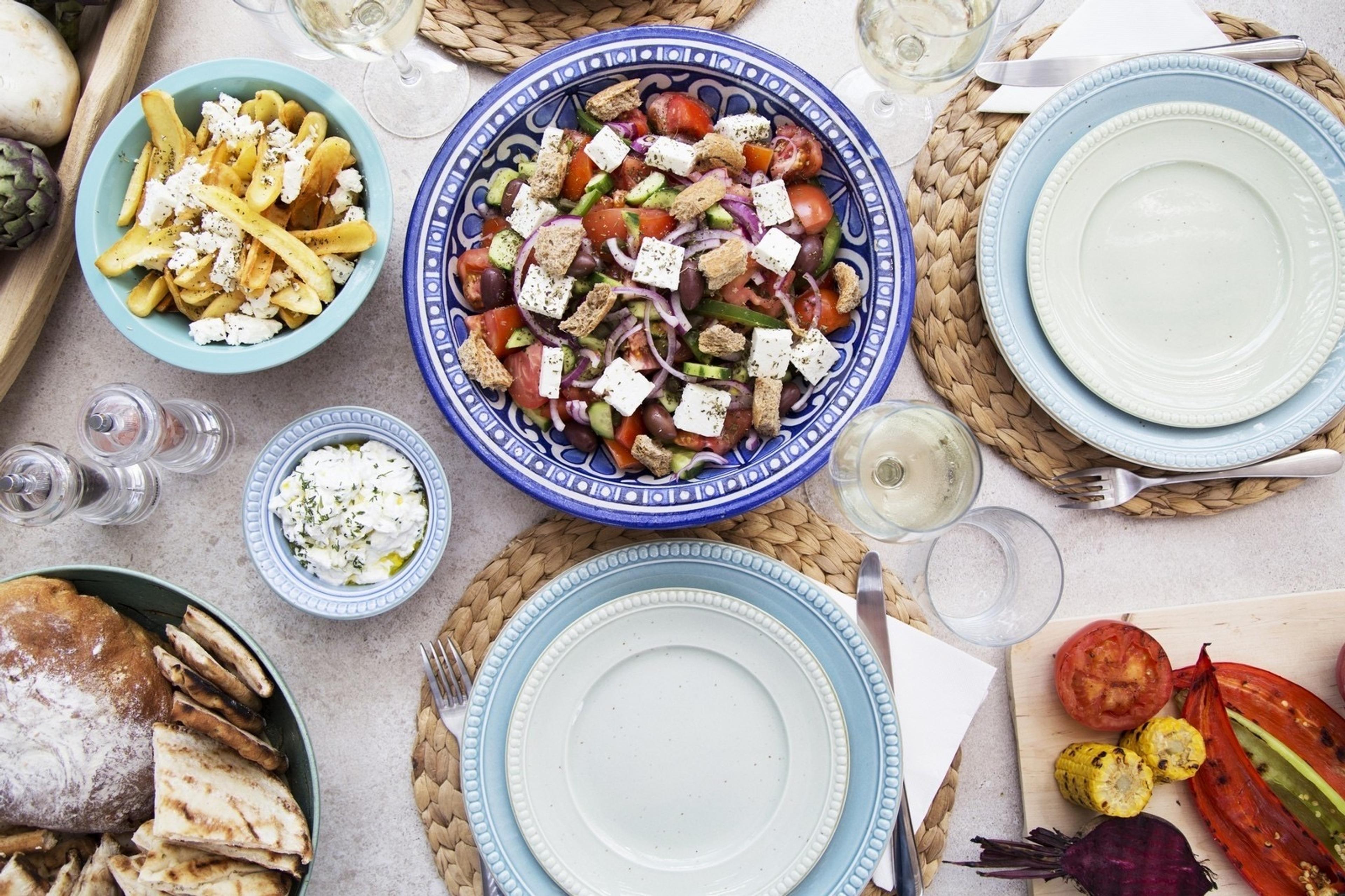 A table laid for several people: a bowl of Greek salad with feta and bread, a bowl of chips and a yoghurt dip, grilled bread, wine glasses and a board of grilled vegetables.