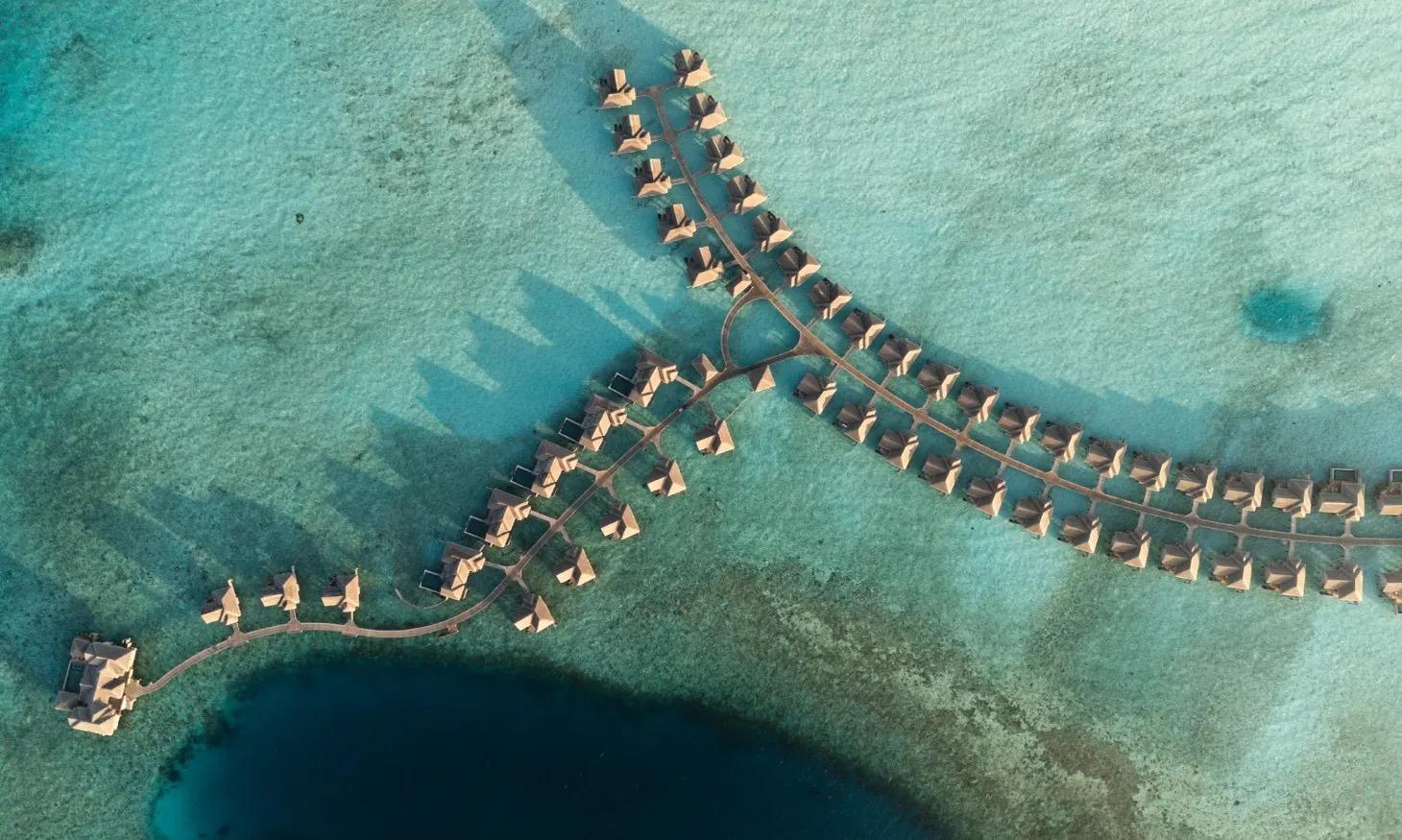 an aerial view of two rows of overwater villas branching out from the island on wooden walkways over pale turquoise reef