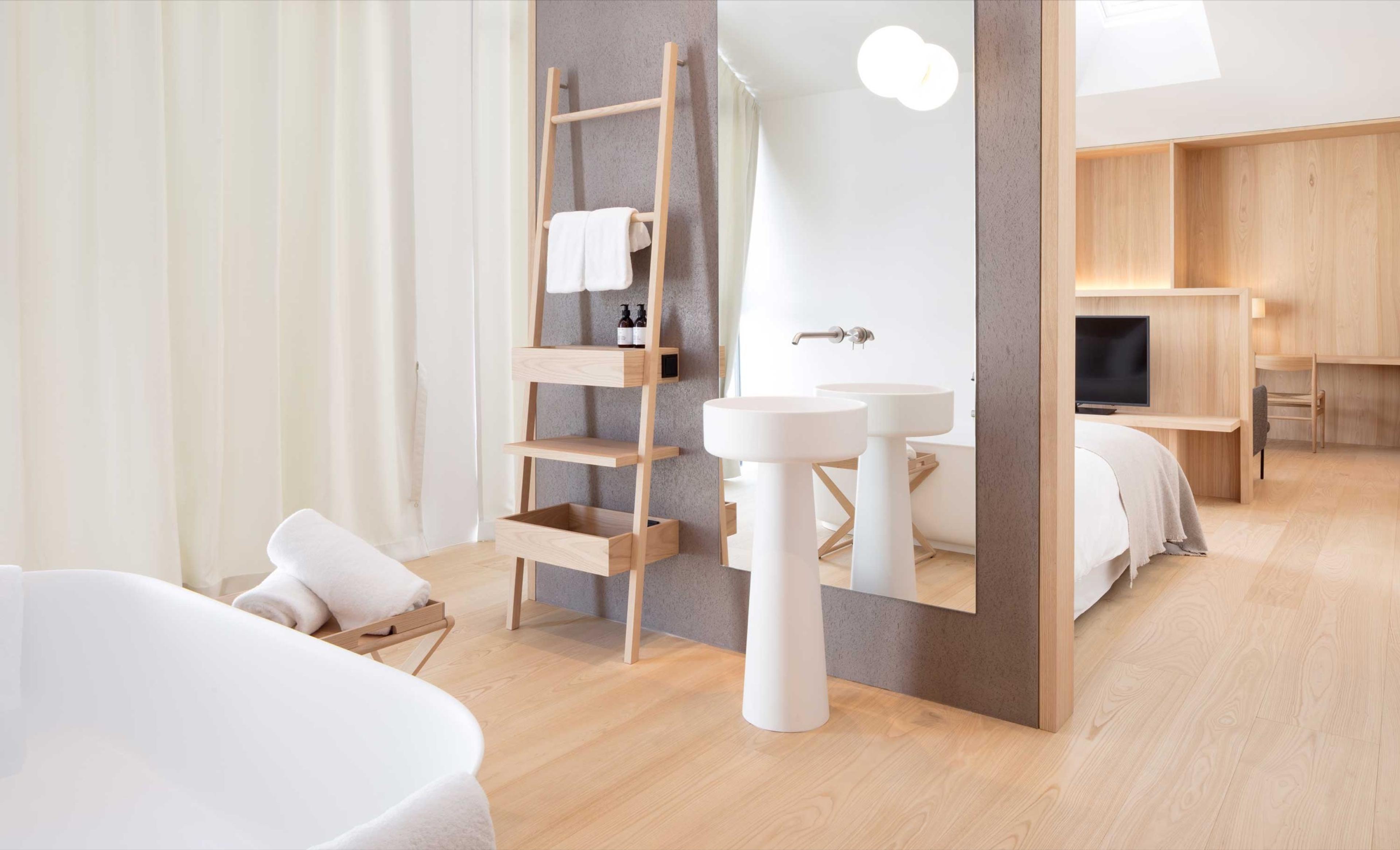 An open hotel bathroom with a freestanding white bathtub, a wooden towel ladder, and two pedestal basins in front of a large mirror reflecting the bed beyond.