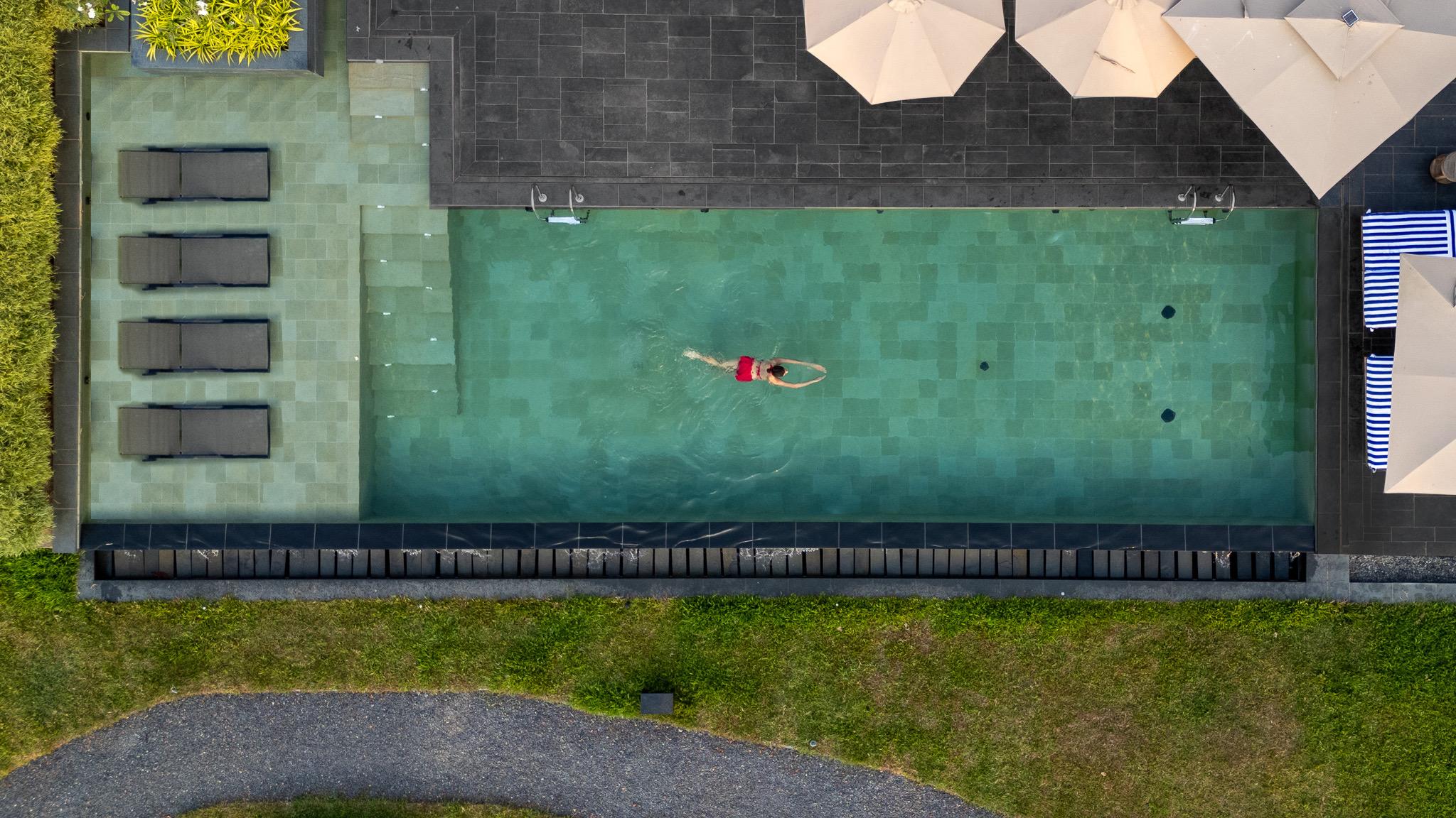 A drone view straight down on a swimming pool with a person swimming in a red swimsuit, sun loungers and umbrellas along one side.