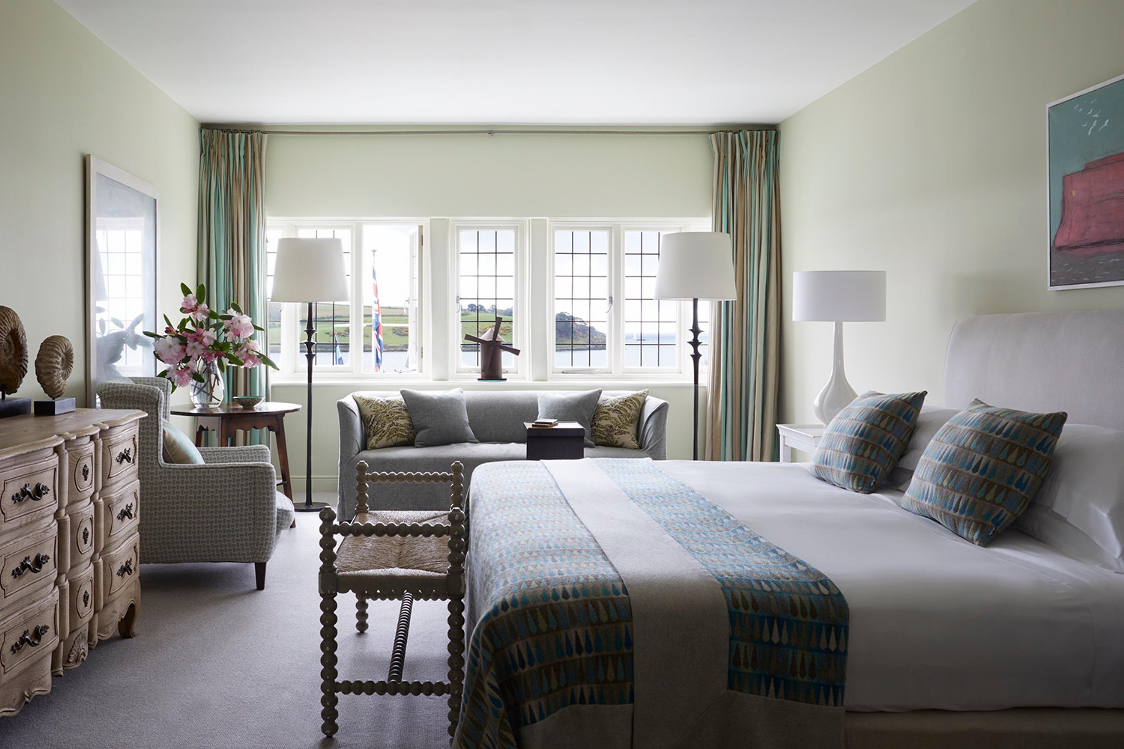 A bedroom with pale green walls, a grey sofa under a row of windows looking onto the water, and a bed dressed in white linen with a patterned throw.