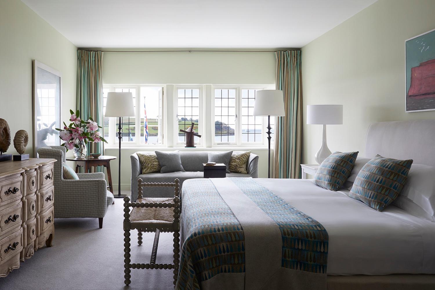A bedroom with pale green walls, a grey sofa under a row of windows looking onto the water, and a bed dressed in white linen with a patterned throw.