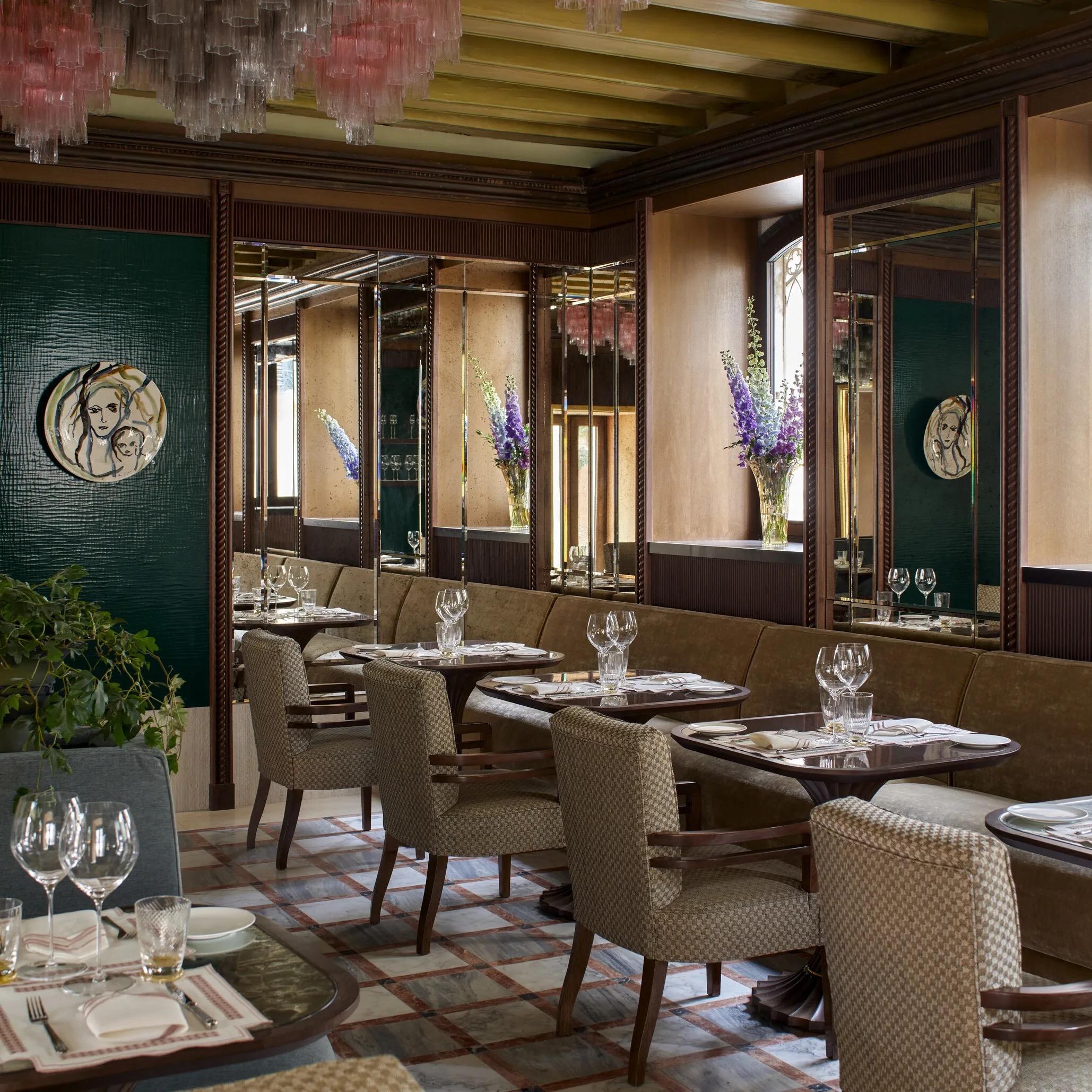 A dining room with pink glass chandeliers, mirrored wood panelling, bench seating, and tables set with wine glasses and a vase of delphiniums