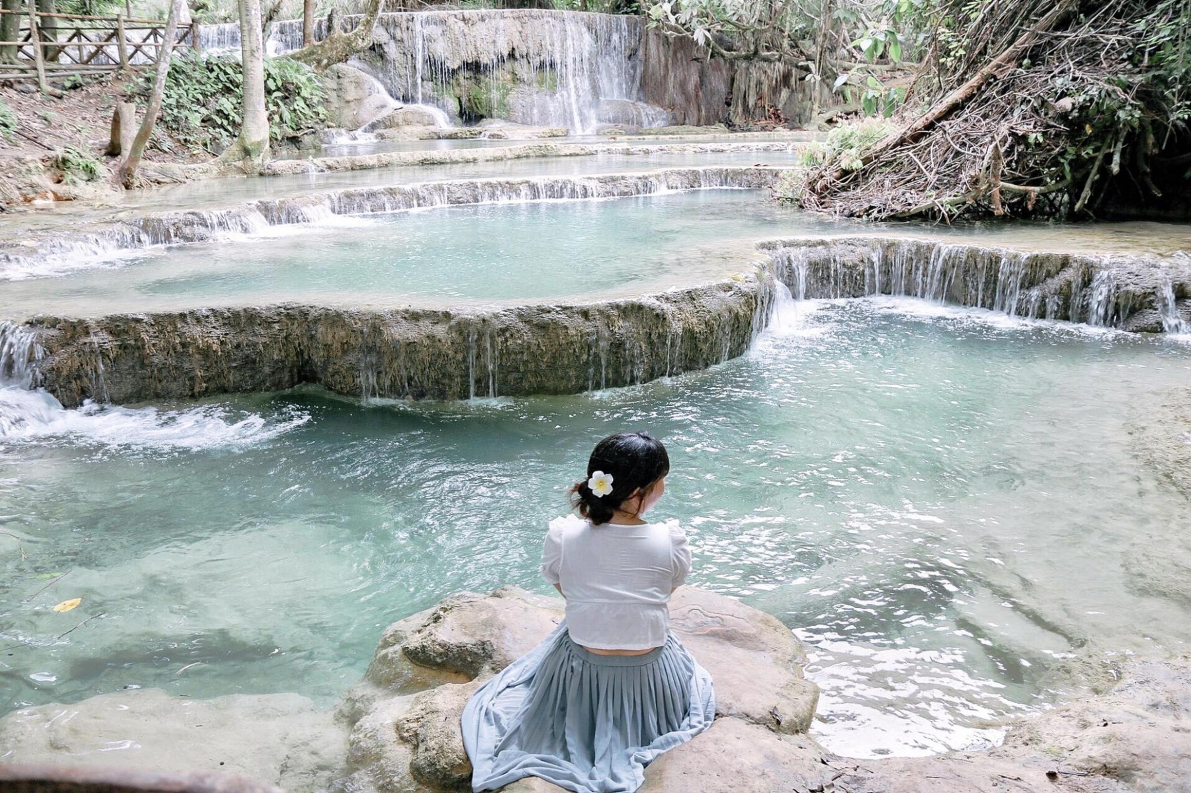 A woman with a flower in her hair sits on a rock looking out over turquoise terraced pools and a small waterfall.