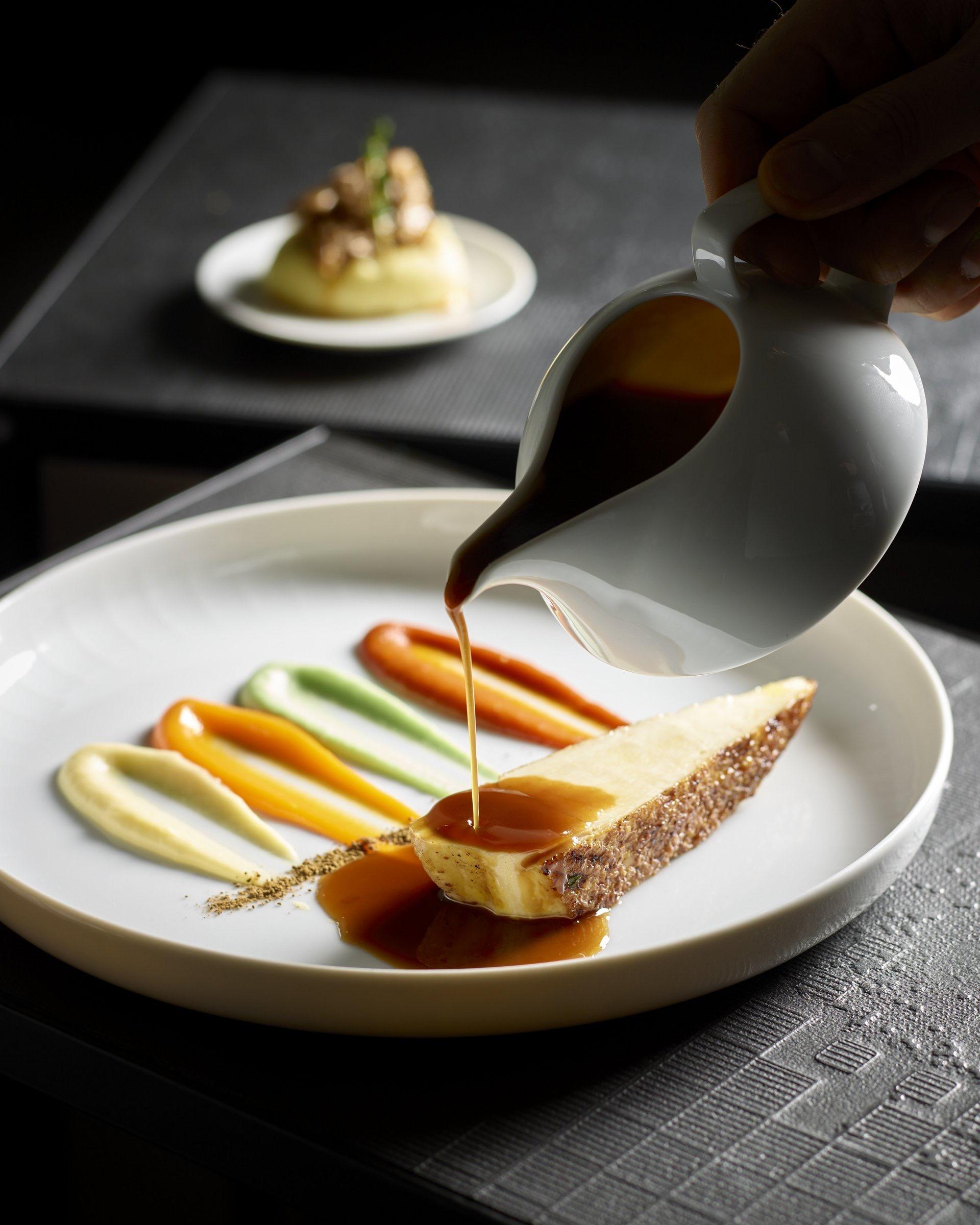a hand pouring sauce from a small jug over a plated dessert, with swipes of coloured puree on the plate