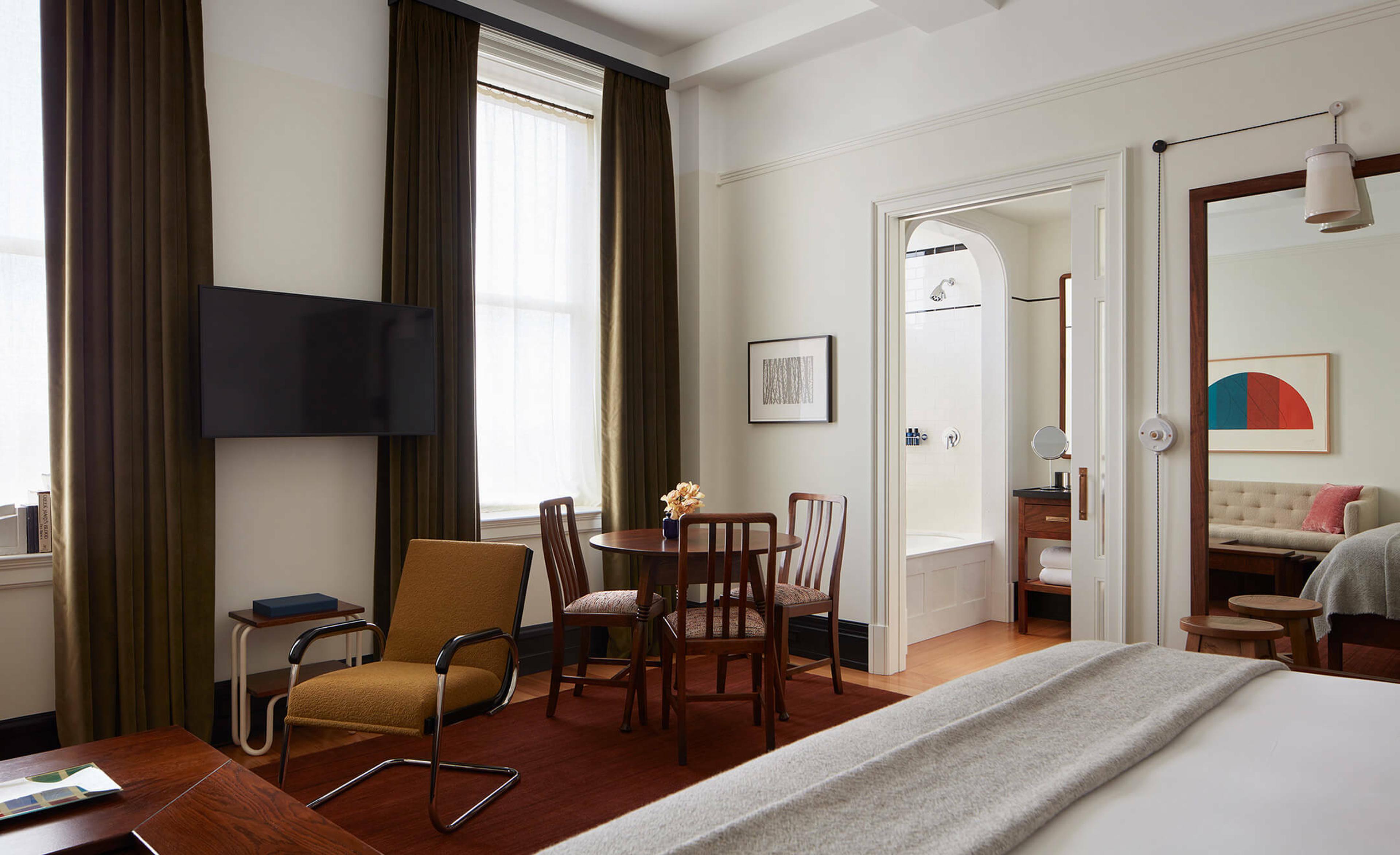 A hotel room with a round table and chairs by curtained windows, a wall-mounted television, an open bathroom door showing a tub and shower, and a tall mirror reflecting a seating area.
