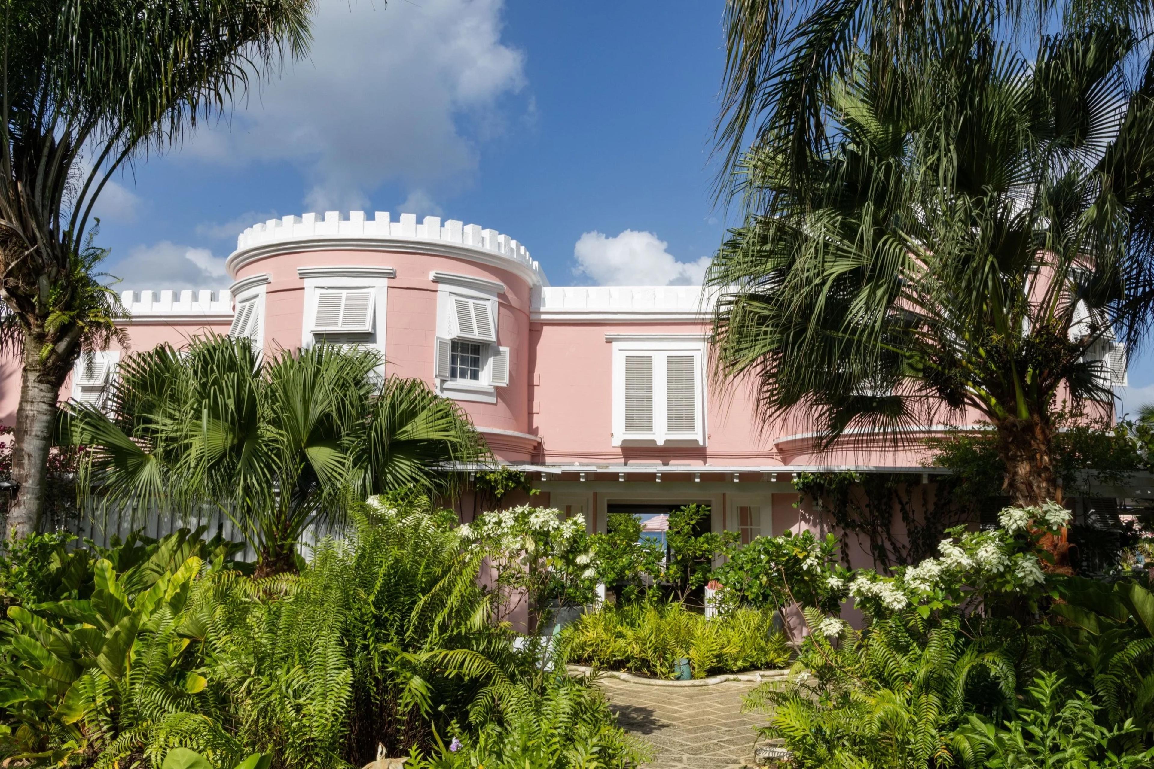 a pink colonial-style building with white crenellated trim, seen through palm trees and tropical garden planting