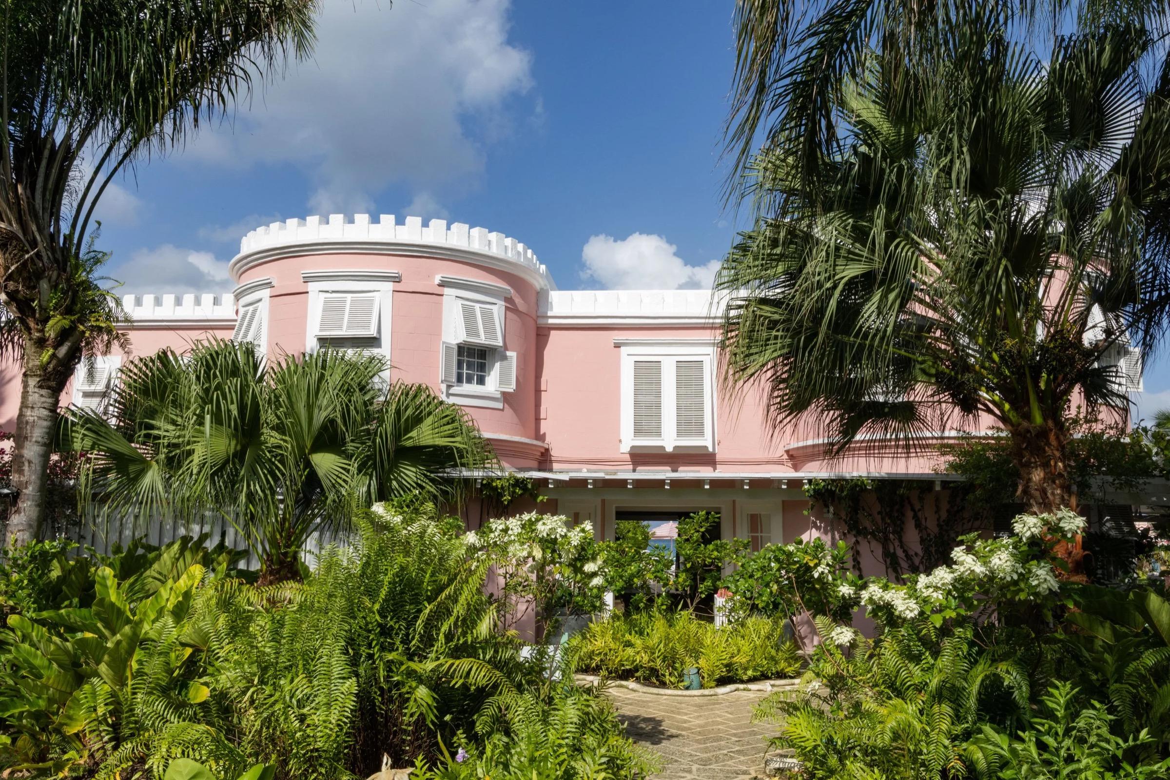a pink colonial-style building with white crenellated trim, seen through palm trees and tropical garden planting