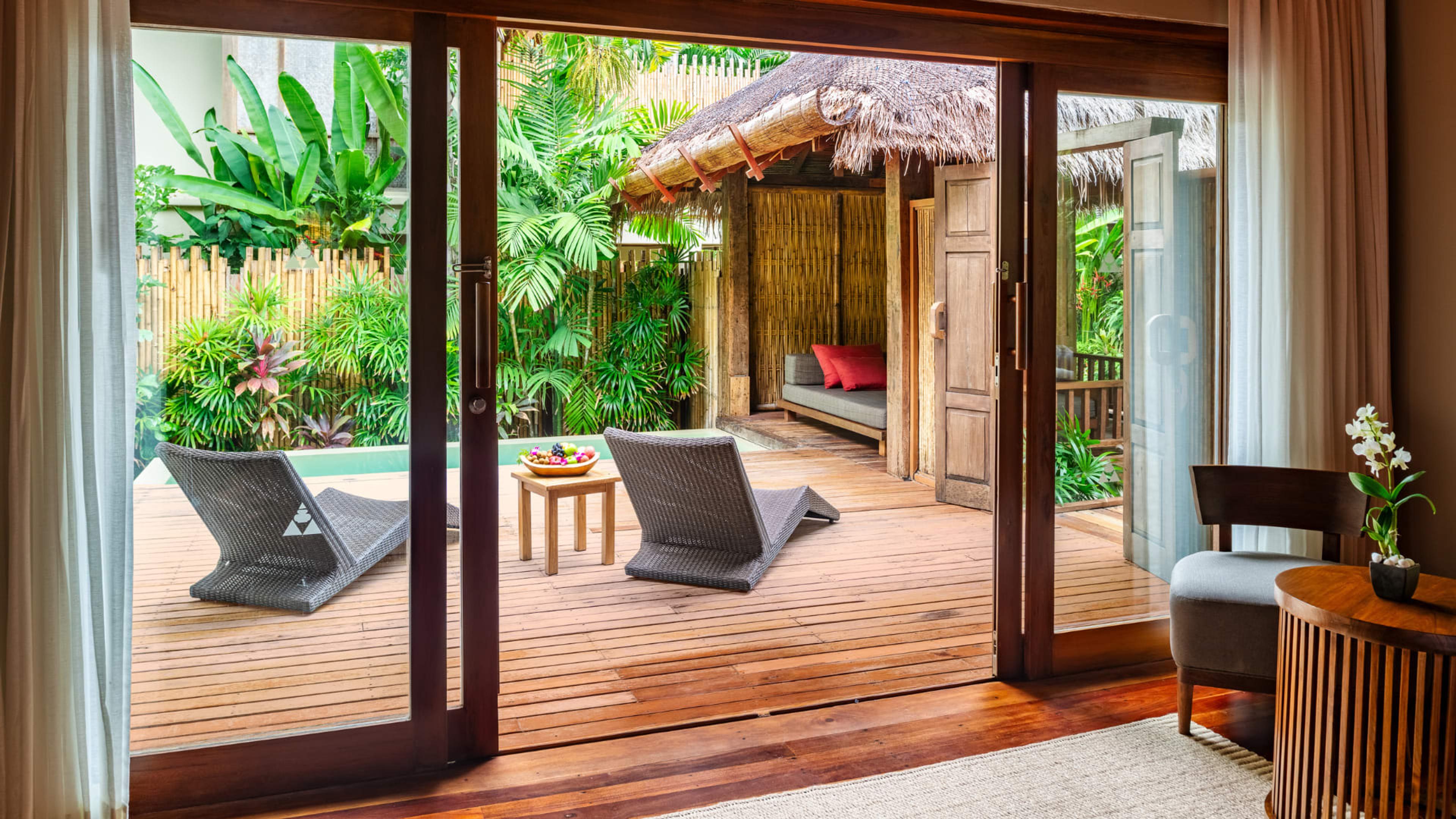 Open sliding doors from a villa's interior onto a wooden deck with two loungers, a small plunge pool and a thatched day bed.