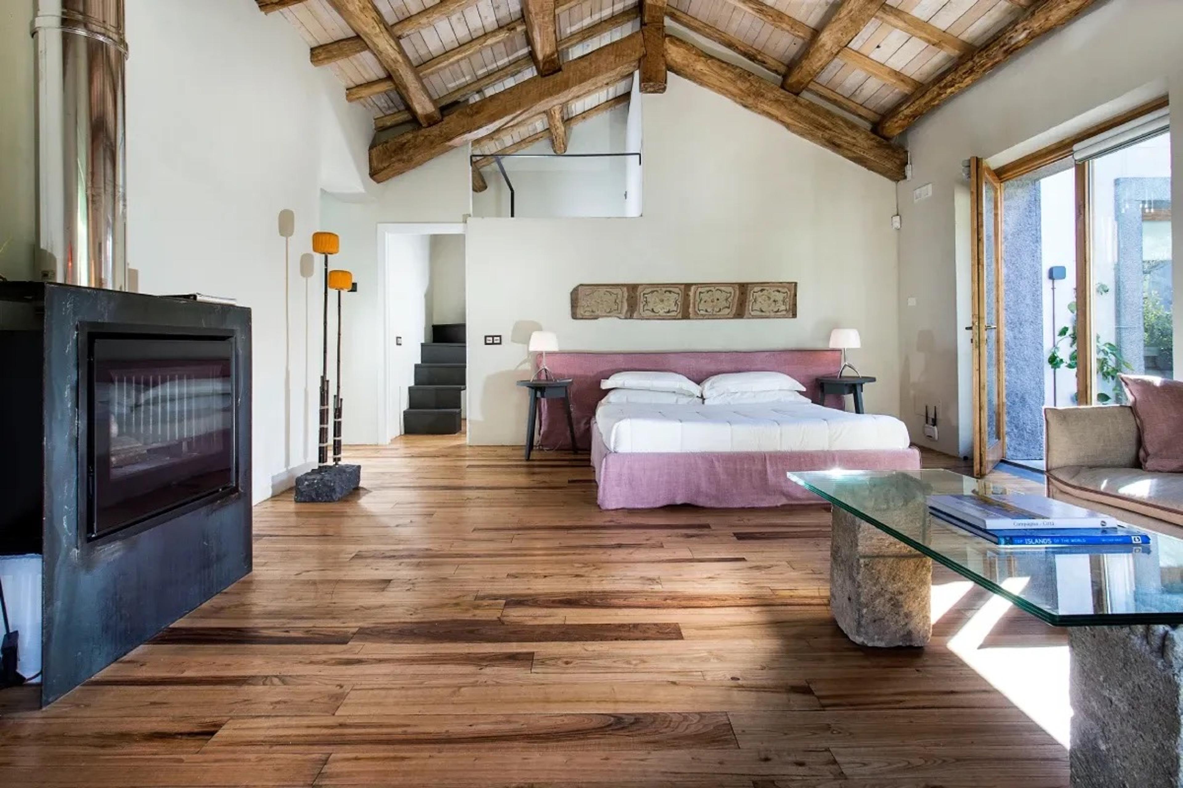 A loft bedroom with a bed, a fireplace, a wood beamed mezzanine above and a glass coffee table with magazines.