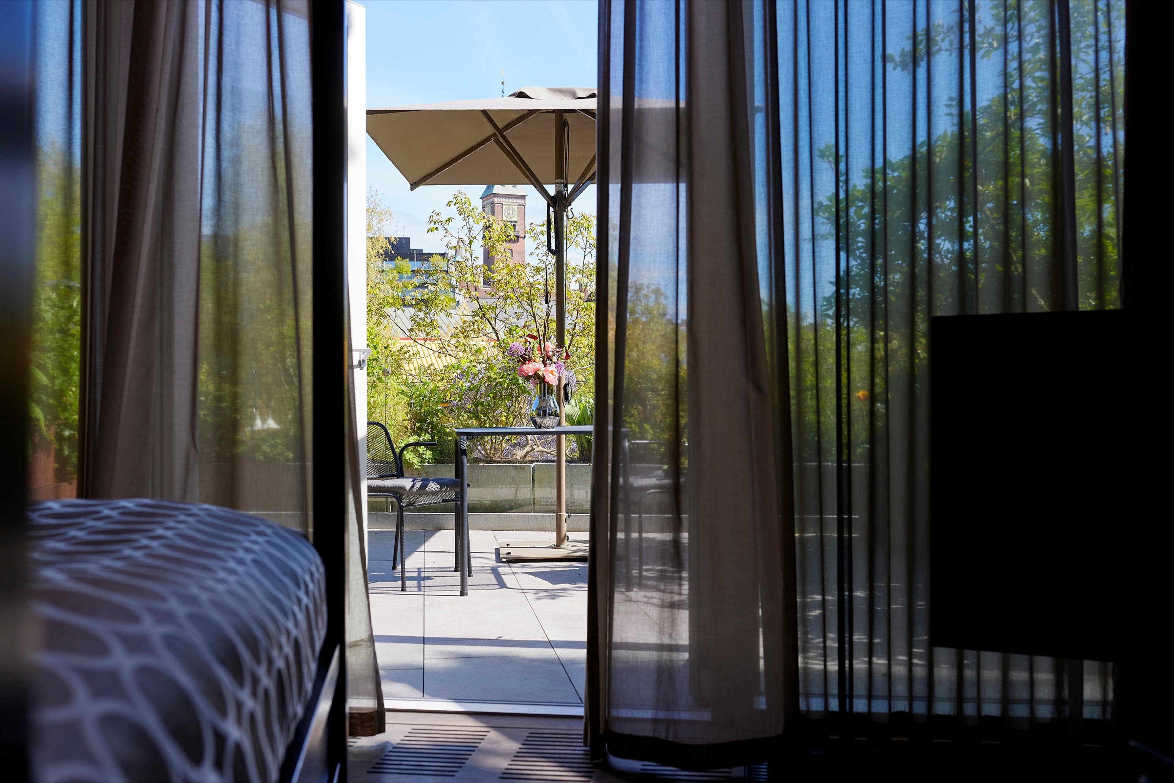 Sheer curtains frame a view through an open balcony door onto a rooftop terrace with a table, chairs and a closed umbrella, trees and a tower beyond.