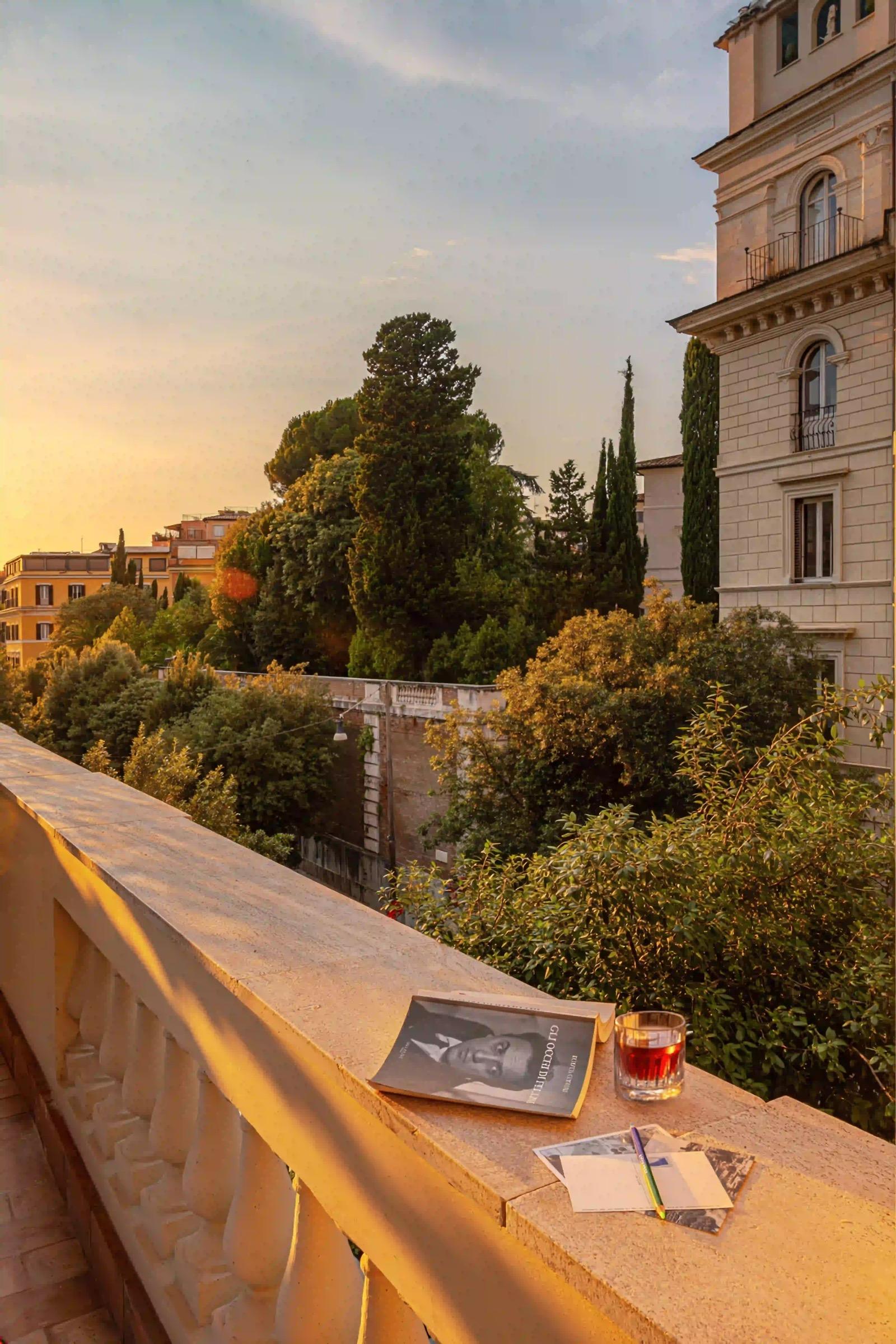 A stone balcony ledge at sunset holding an open book, a glass of a dark drink, a pencil and postcards, overlooking trees and rooftops.