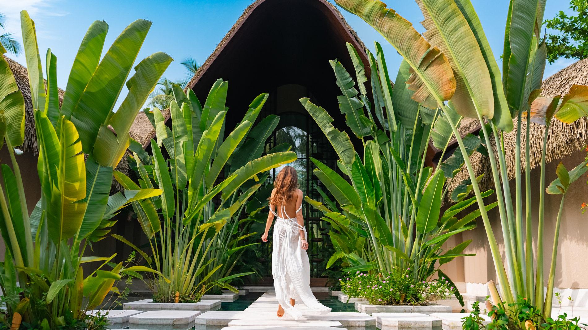 A woman walking away from the camera along a stone path through tall banana leaves toward a dark thatched entrance.